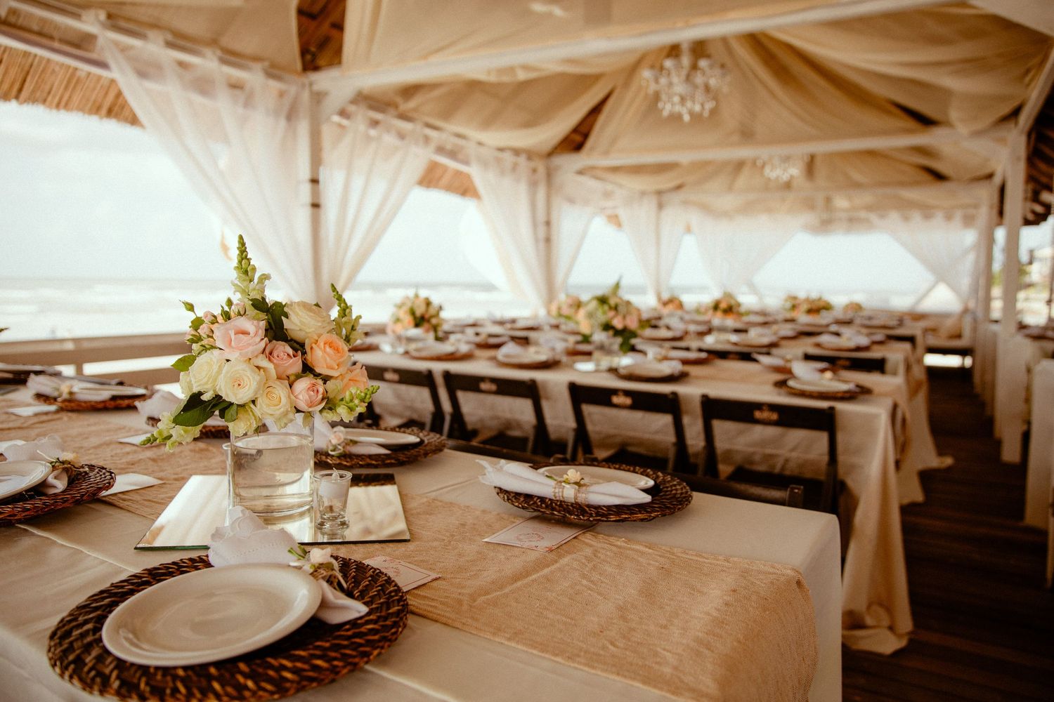 Elegant wedding reception table settings with wooden centerpieces and white tablecloths in a light-filled tent venue.