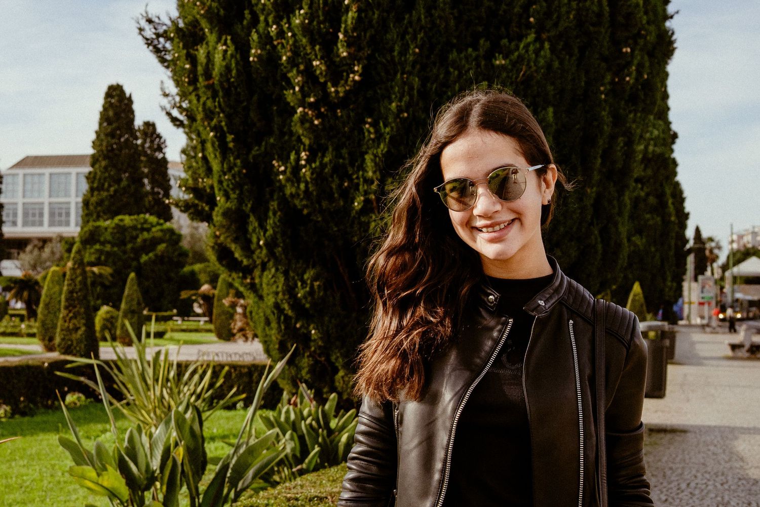 Candid portrait of someone in a black leather jacket smiling outdoors with cypress trees in background.
