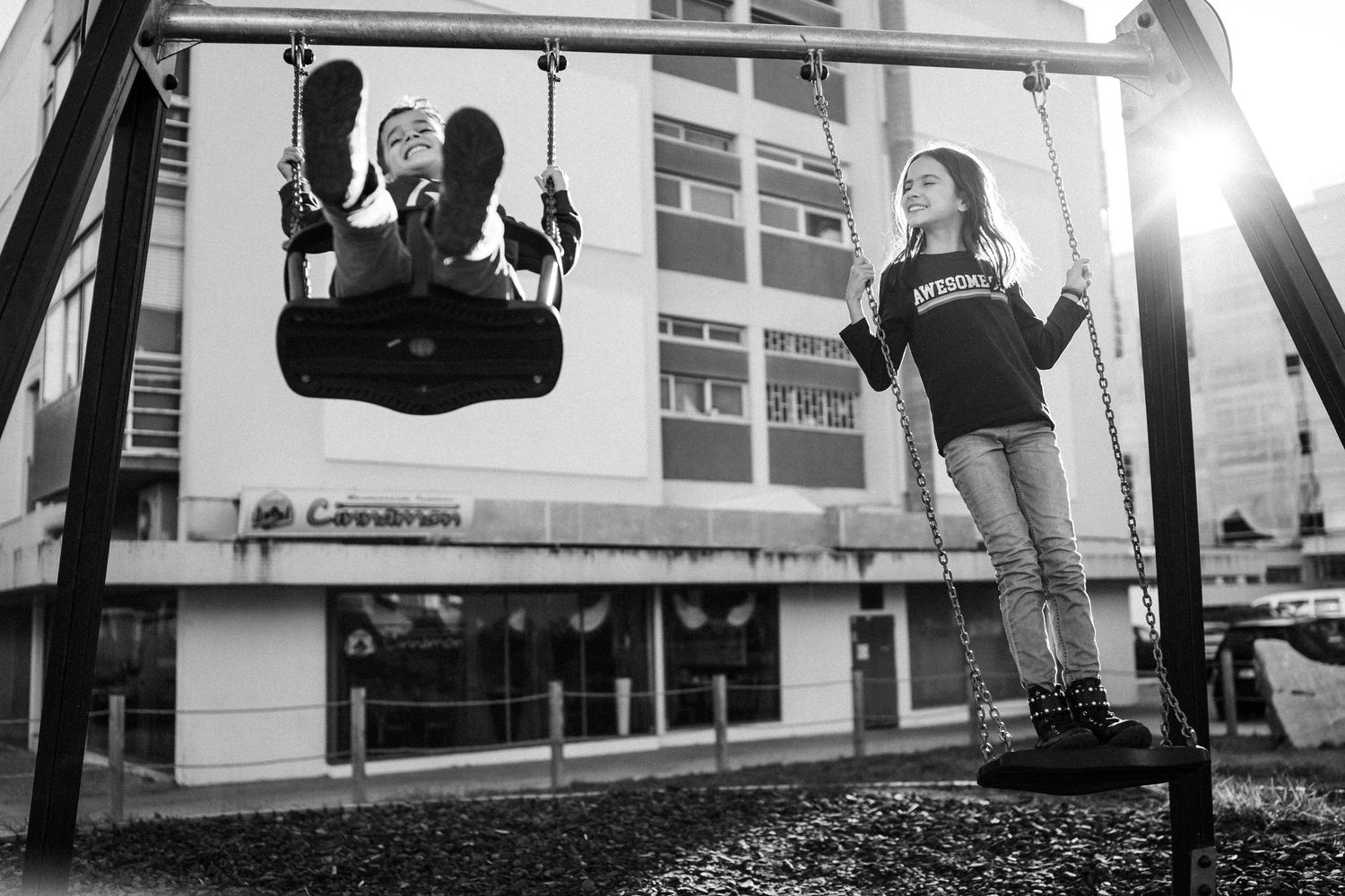 Two people enjoying swings at a playground with sunlight streaming through in black and white photography.