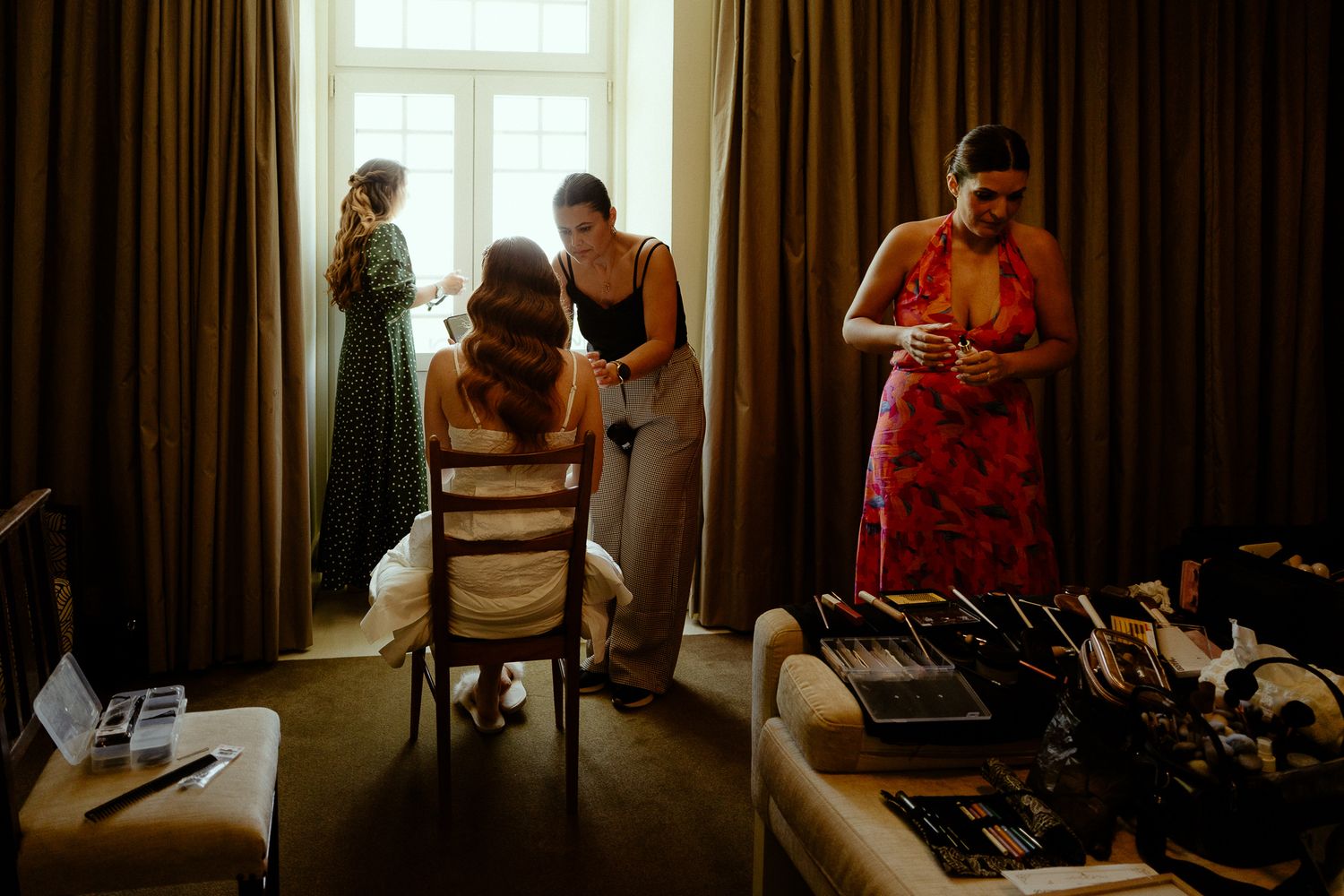 Bridesmaids preparing in a hotel room, with makeup tools on a table and elegant dresses near bright windows.