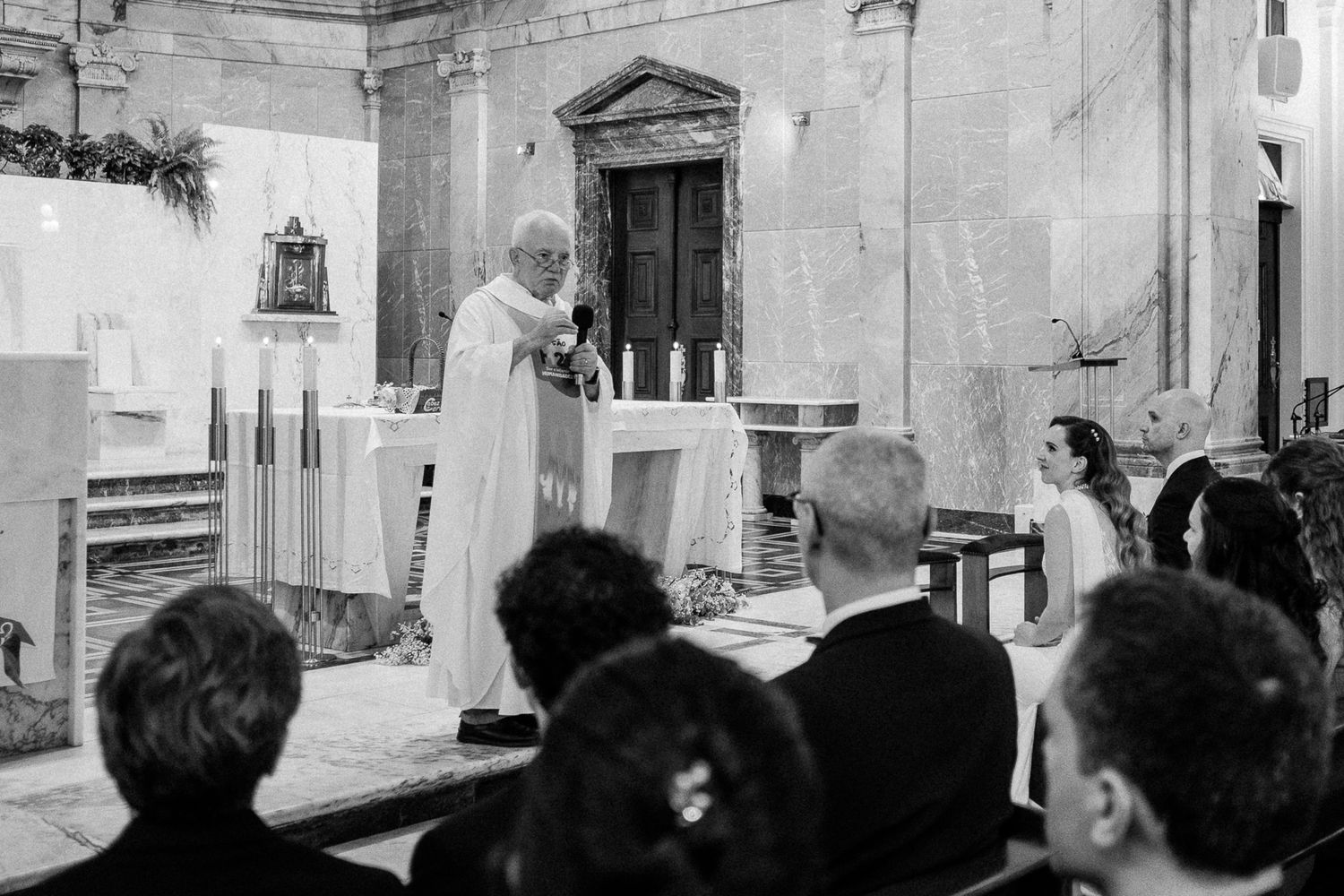 Black and white photo of a priest officiating a wedding ceremony at a church altar with candles and guests seated.