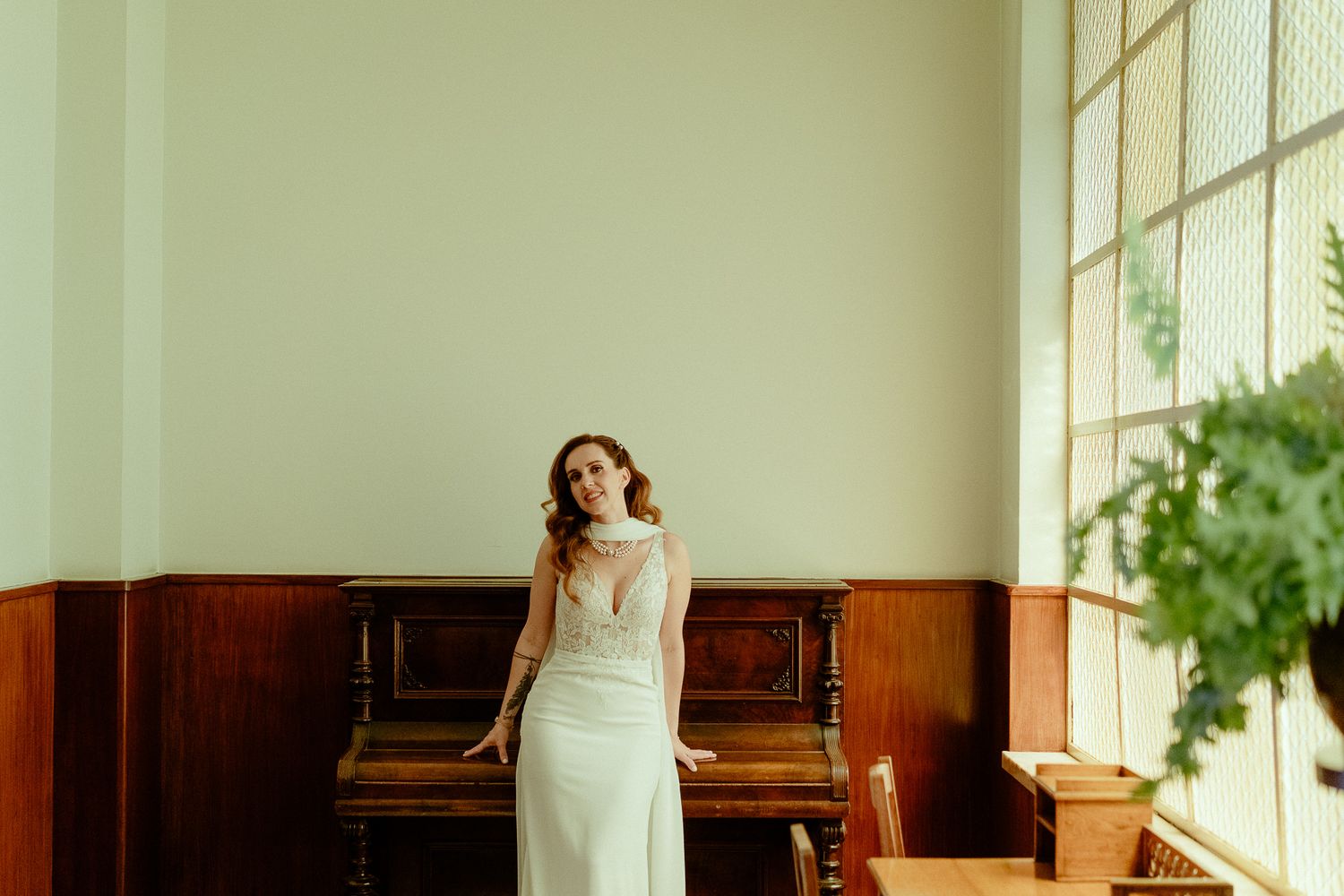 Bride in elegant white gown standing near vintage piano by large window in bright room.