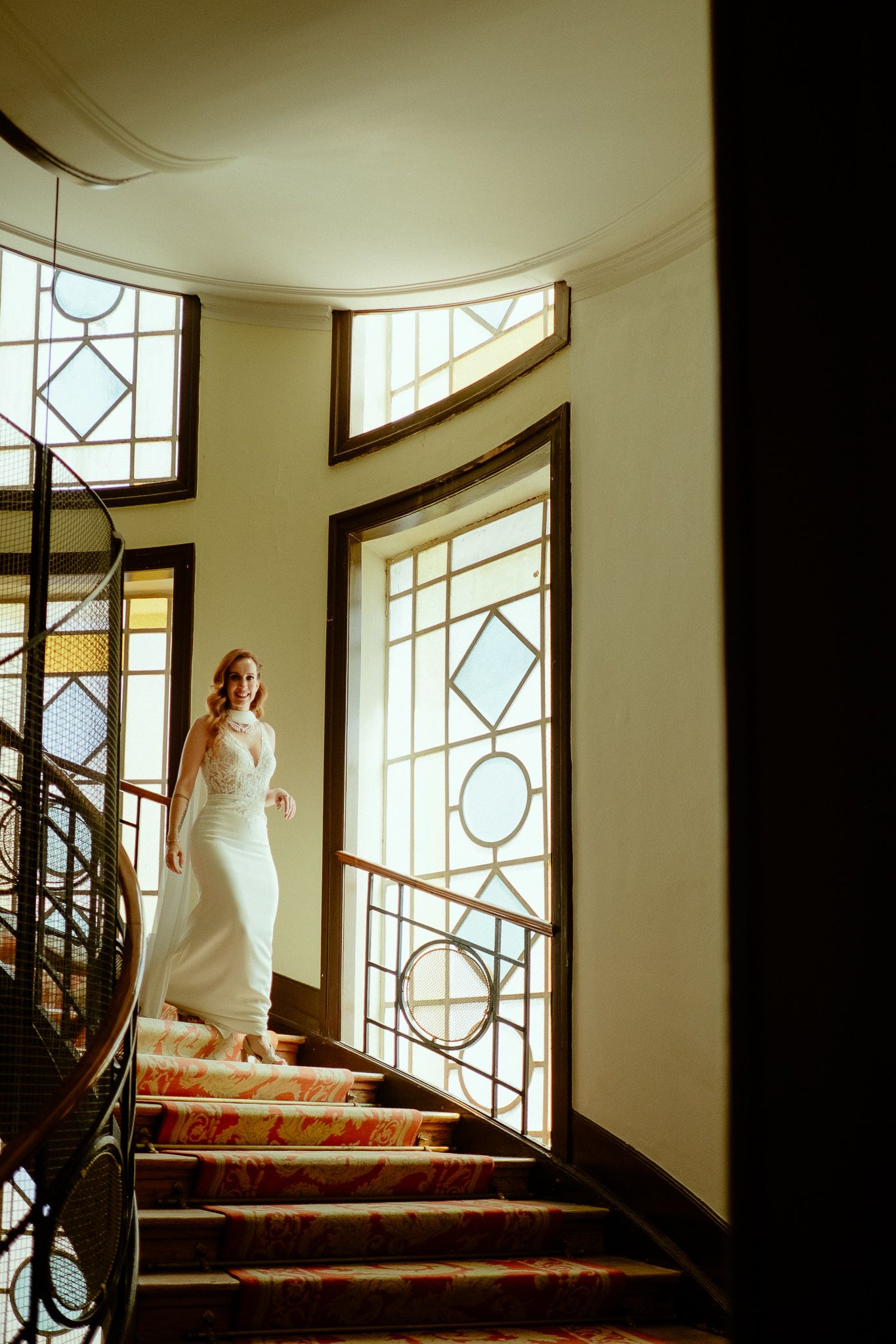 Bride in sleek white halter gown posing on curved staircase with stained glass windows in grand interior.