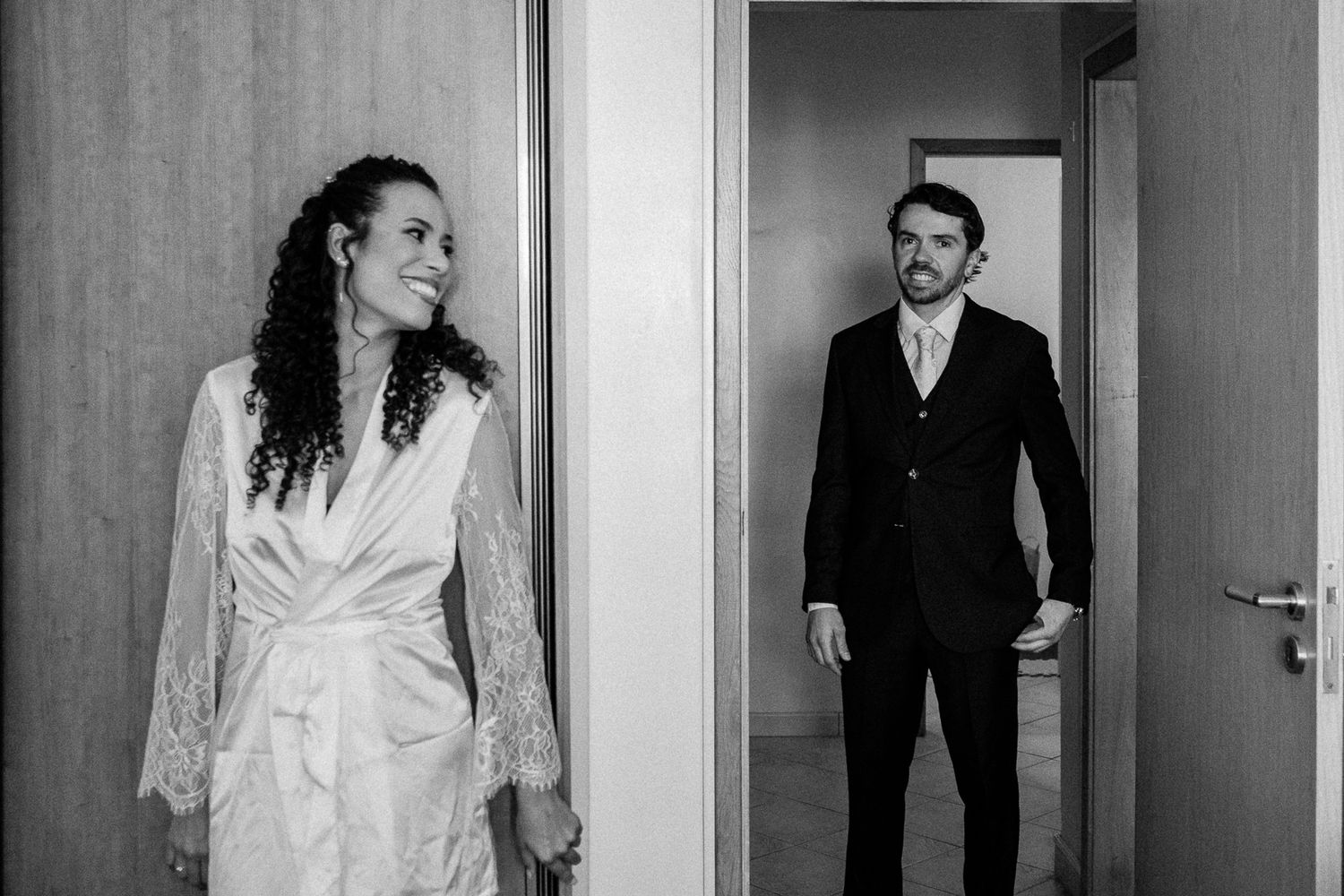 Black and white photo of a smiling bride in lace robe and groom in suit standing in doorways.