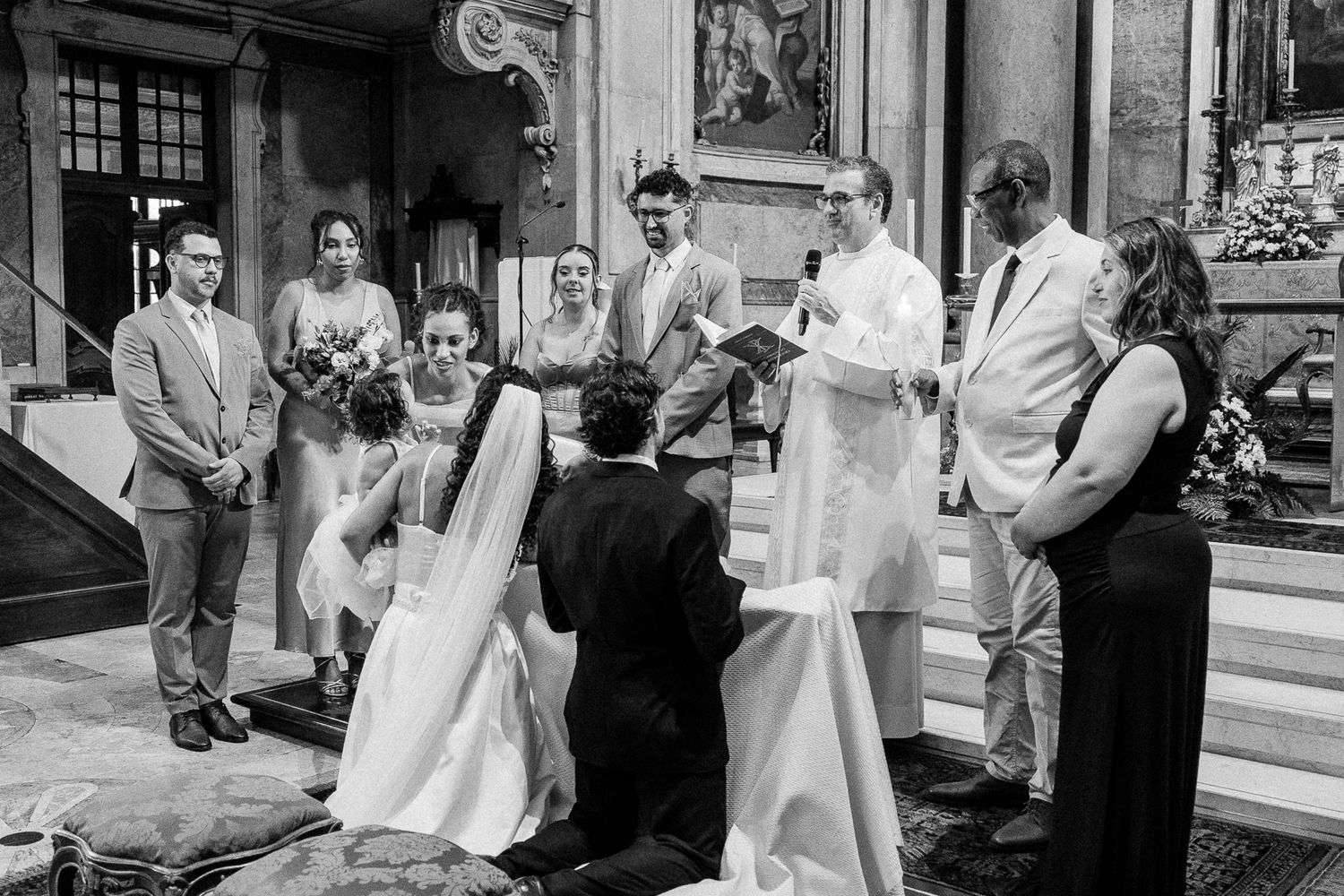 Black and white photo of a wedding ceremony in an ornate church with bride, groom kneeling, officiant, and guests.