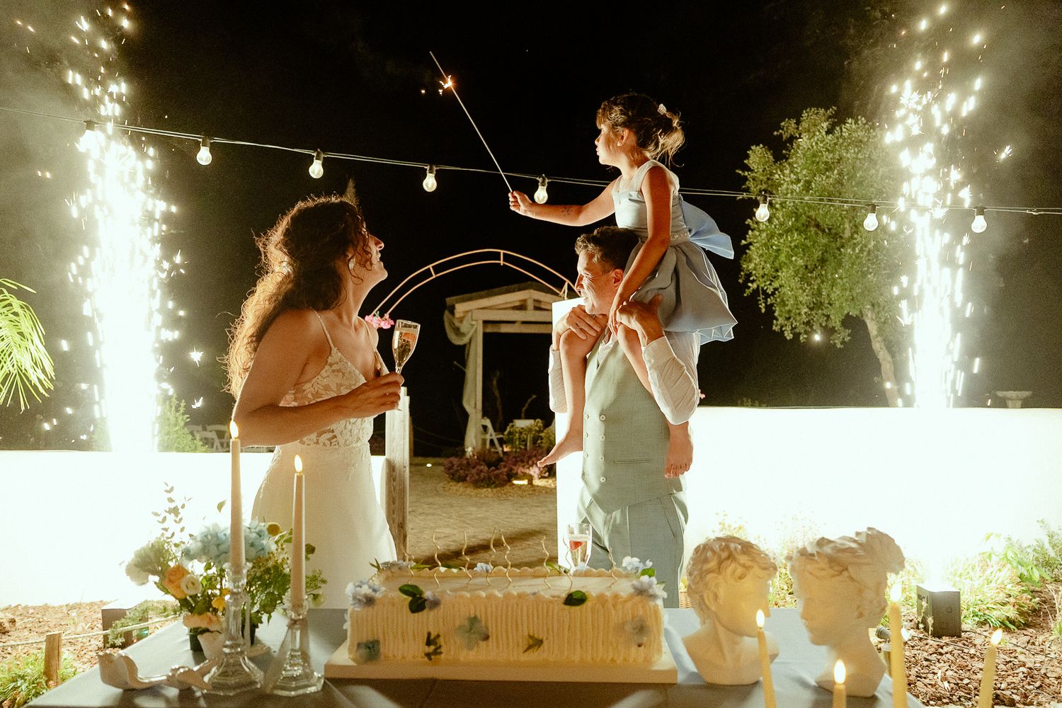 Family celebrating with sparklers beside a wedding cake outdoors at night with festive string lights.