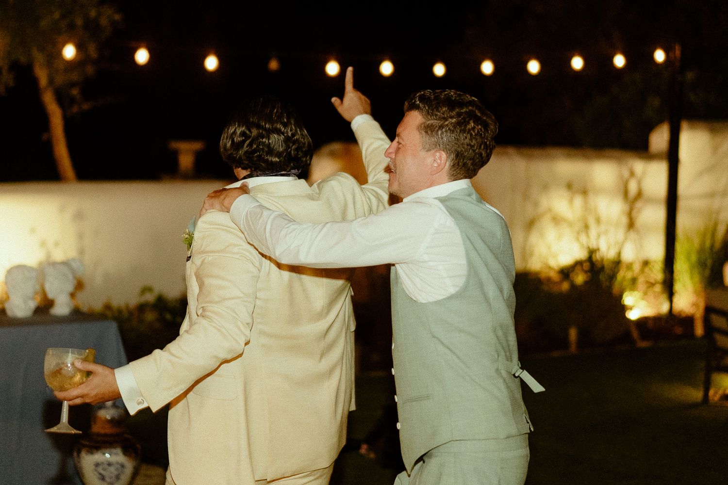 Two grooms embracing and celebrating at an outdoor wedding reception with string lights at night.