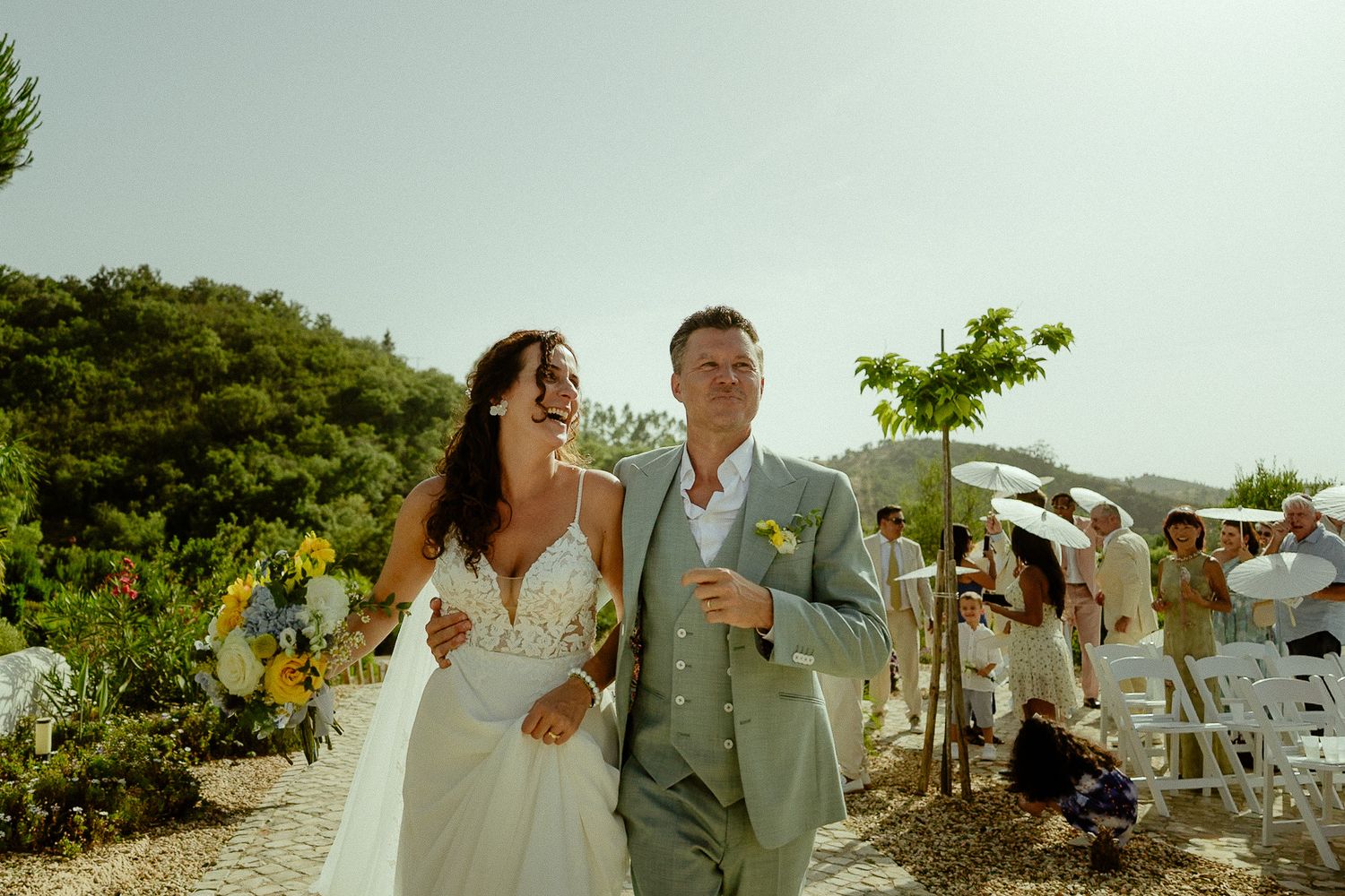Happy newlyweds walk down the aisle at an outdoor wedding ceremony on a sunny hillside.
