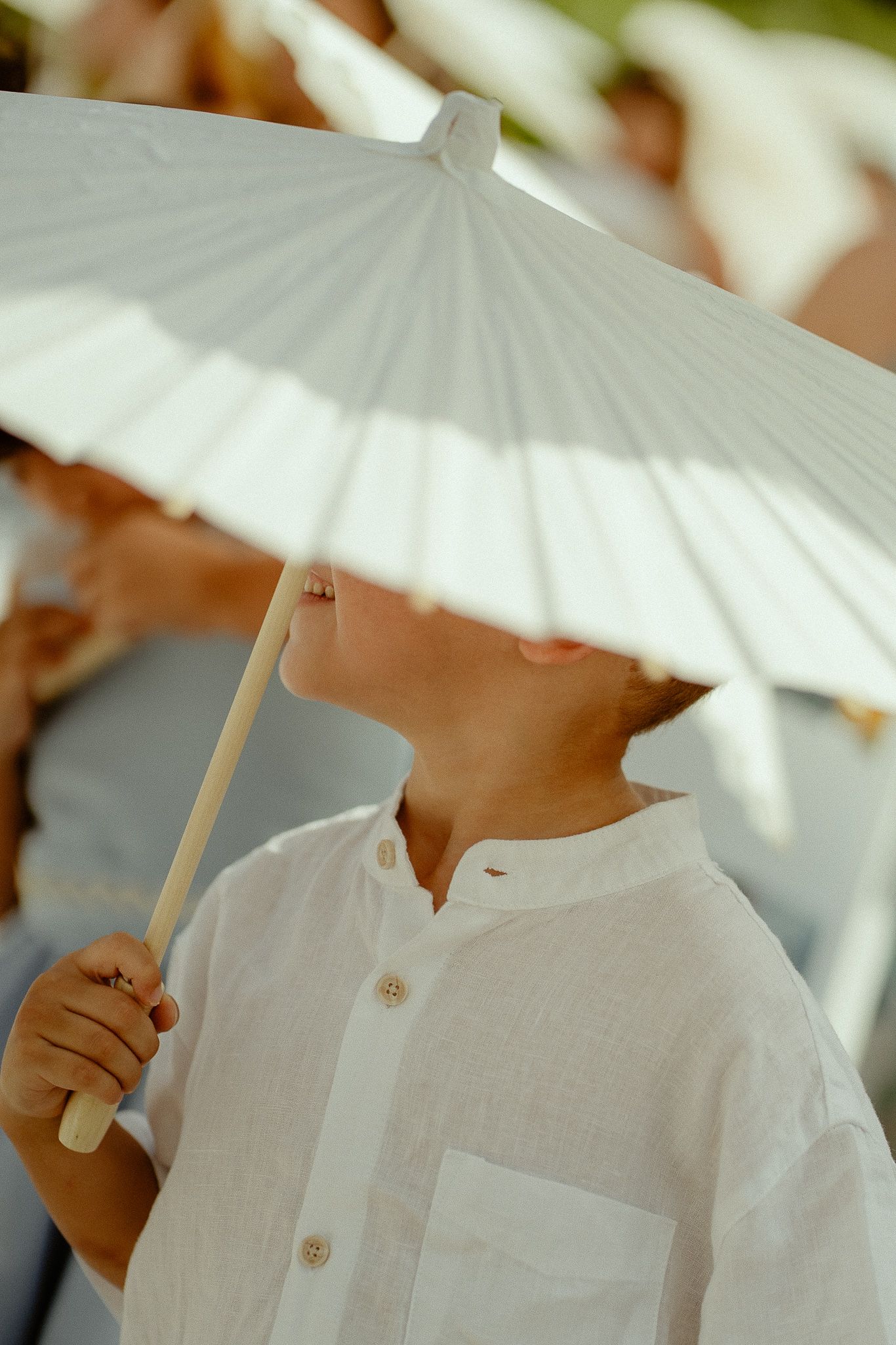 Young boy in white linen shirt holding a white paper parasol umbrella outdoors at a sunny event.
