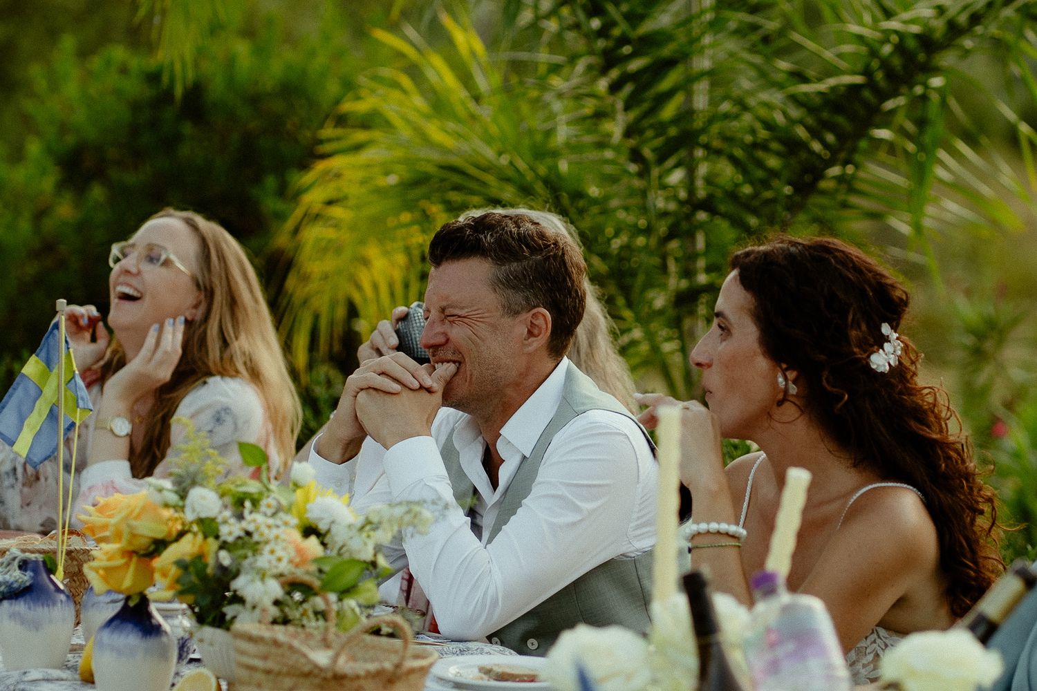 Guests laughing and celebrating at an outdoor wedding reception table surrounded by lush greenery and floral arrangements.
