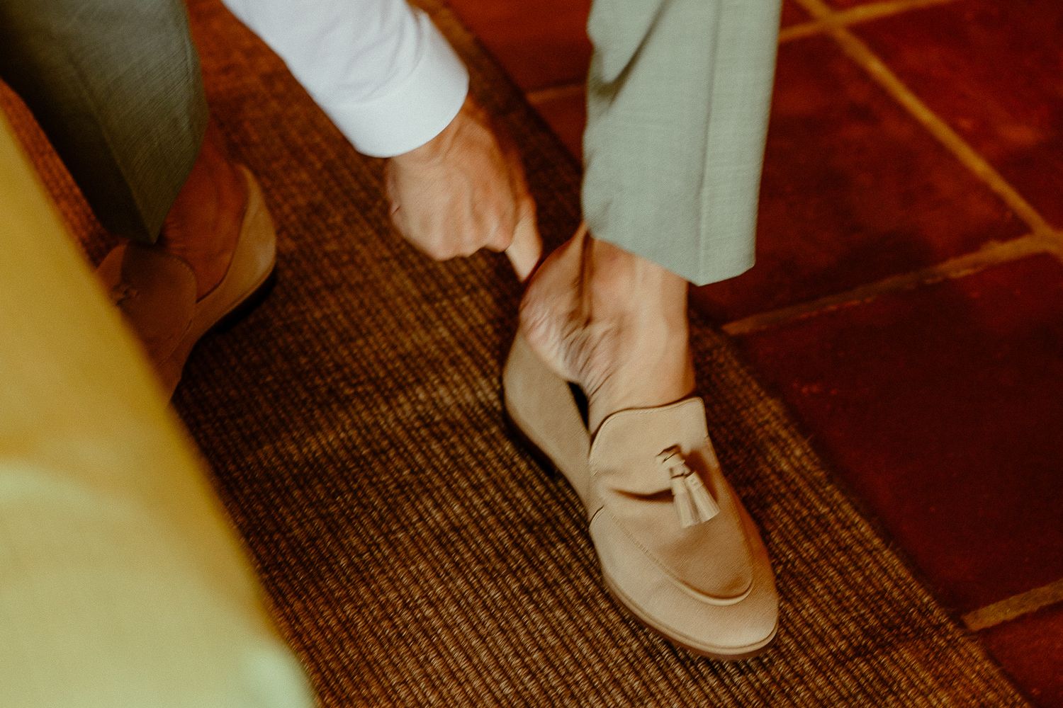 Groom putting on beige loafer shoes with buckle detail on brown carpet before wedding ceremony.