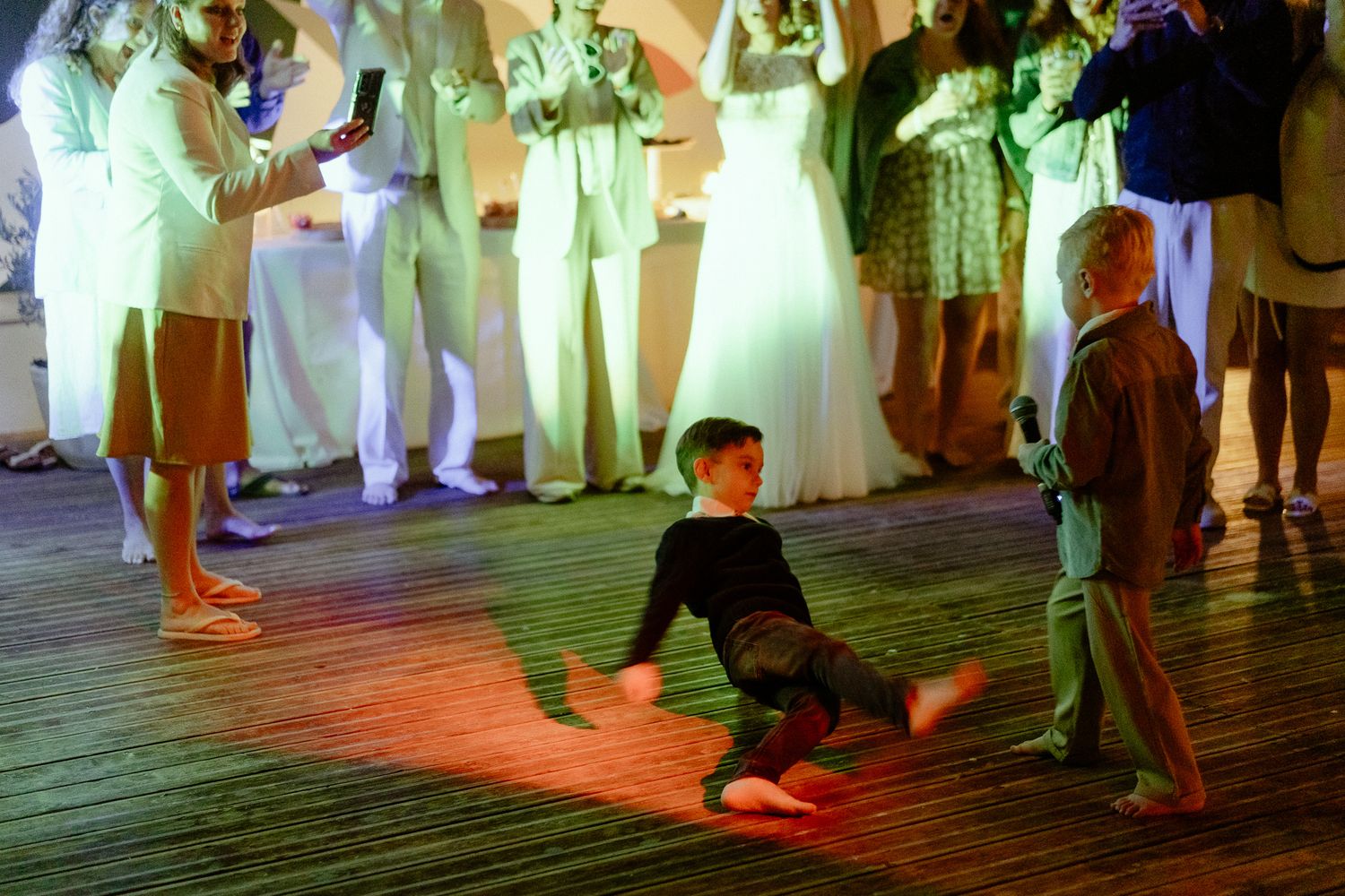 Children dancing energetically on a wooden dance floor at a wedding reception with guests watching in background.