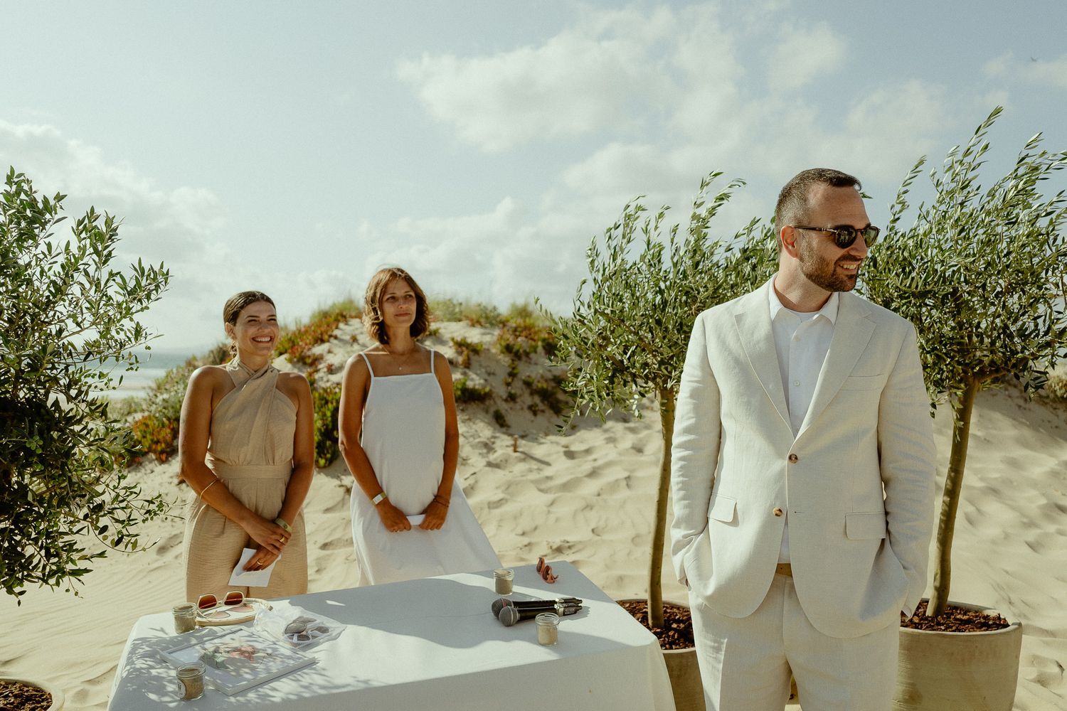 Elegant beach wedding ceremony with groom in white suit and guests standing by a decorated table on sandy dunes.