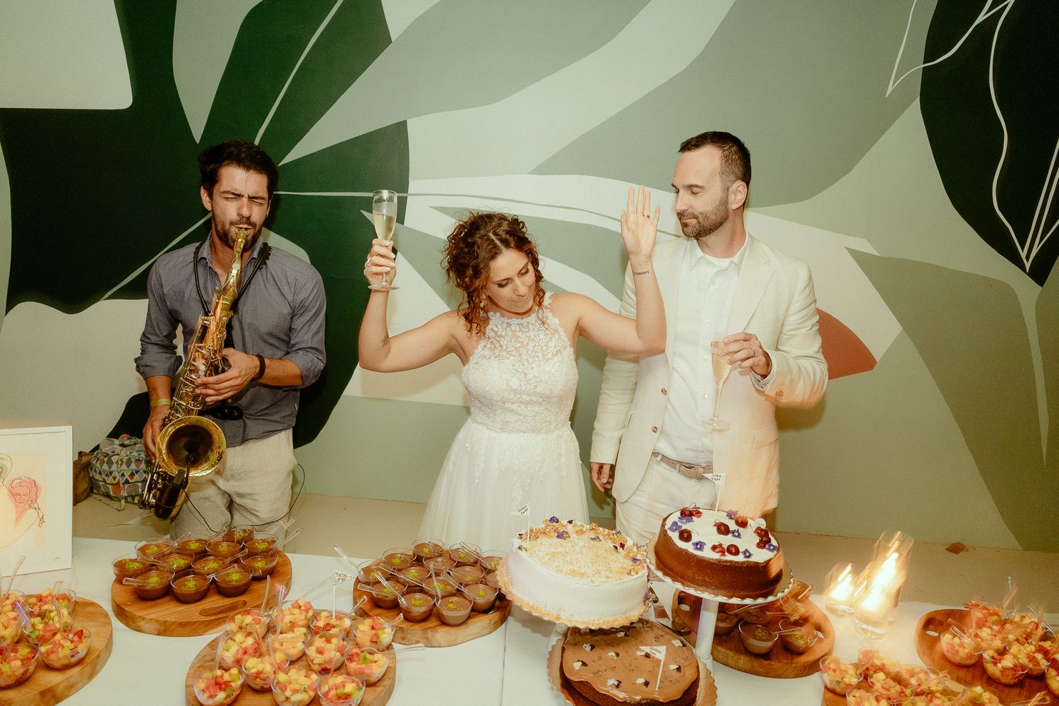 Bride and groom cutting wedding cake while saxophonist performs, surrounded by desserts and candles.
