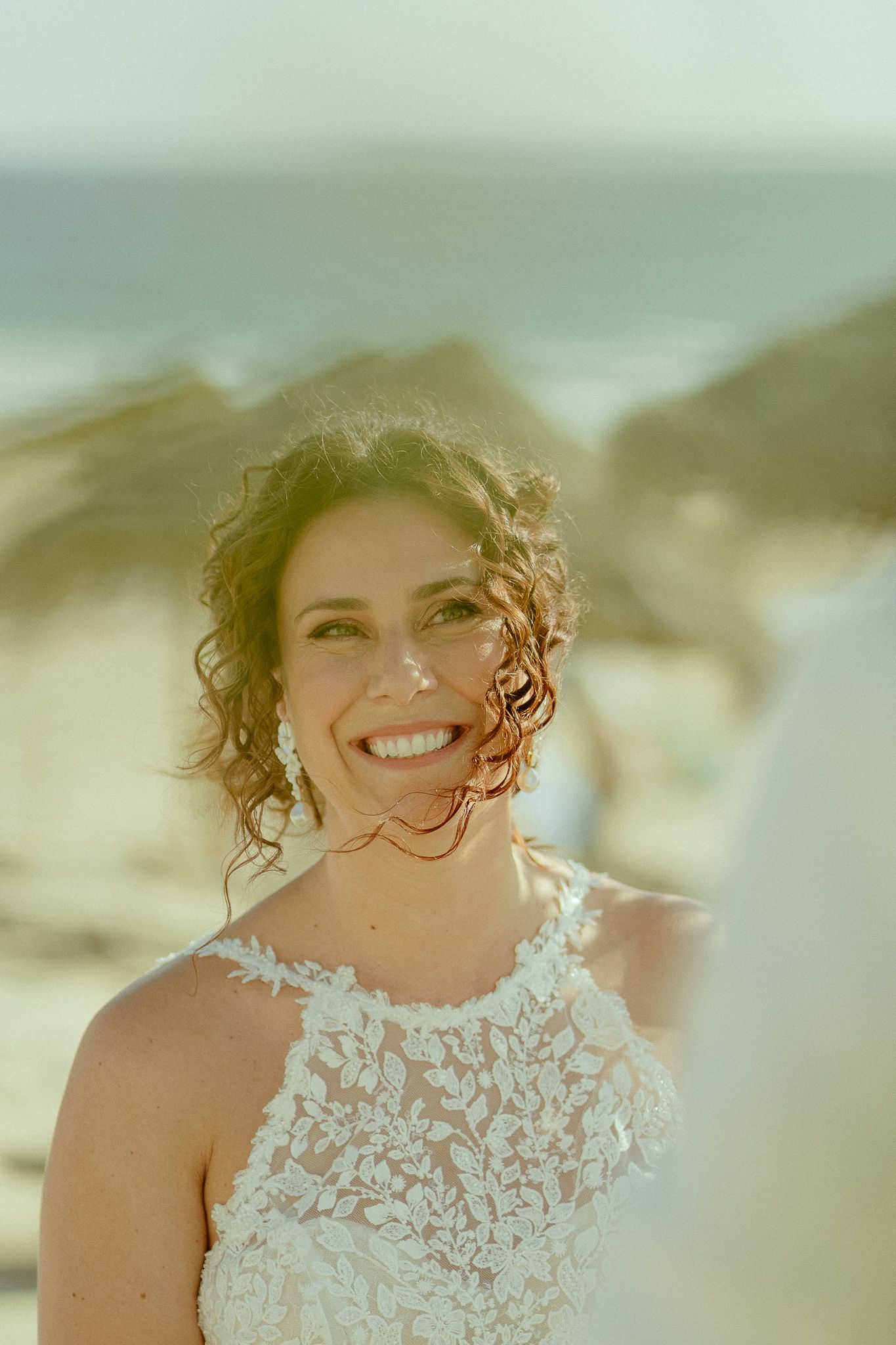 Smiling bride in lace halter wedding dress at beach ceremony with ocean backdrop and warm sunlight.