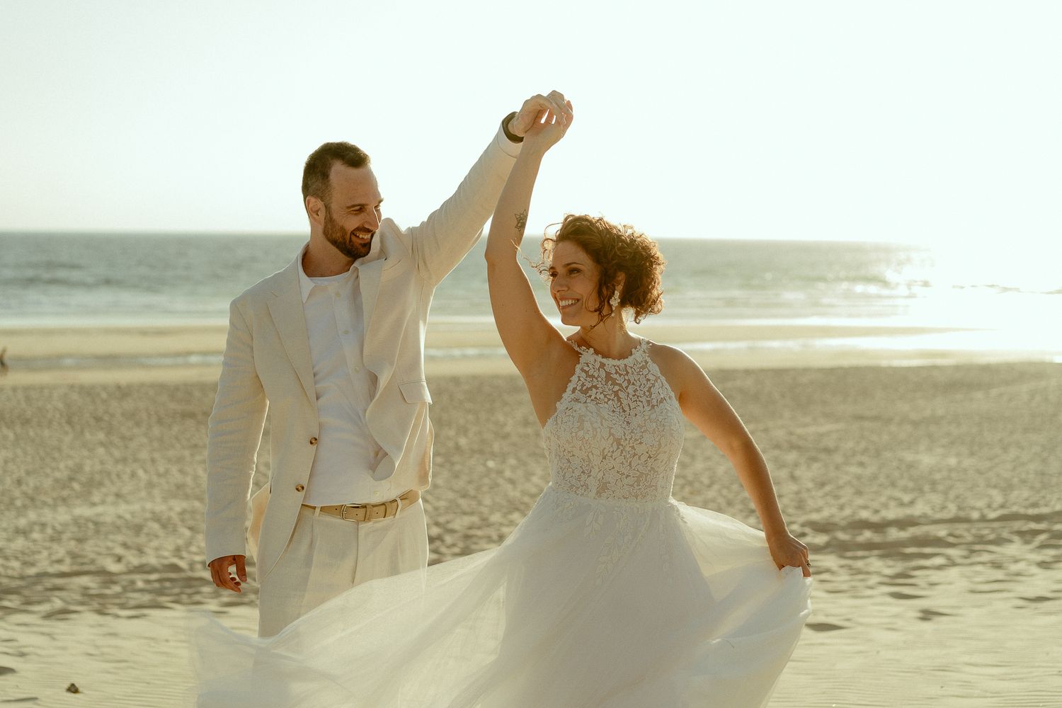 Bride and groom dancing on a sunny beach, bride in white lace gown, groom in cream suit.