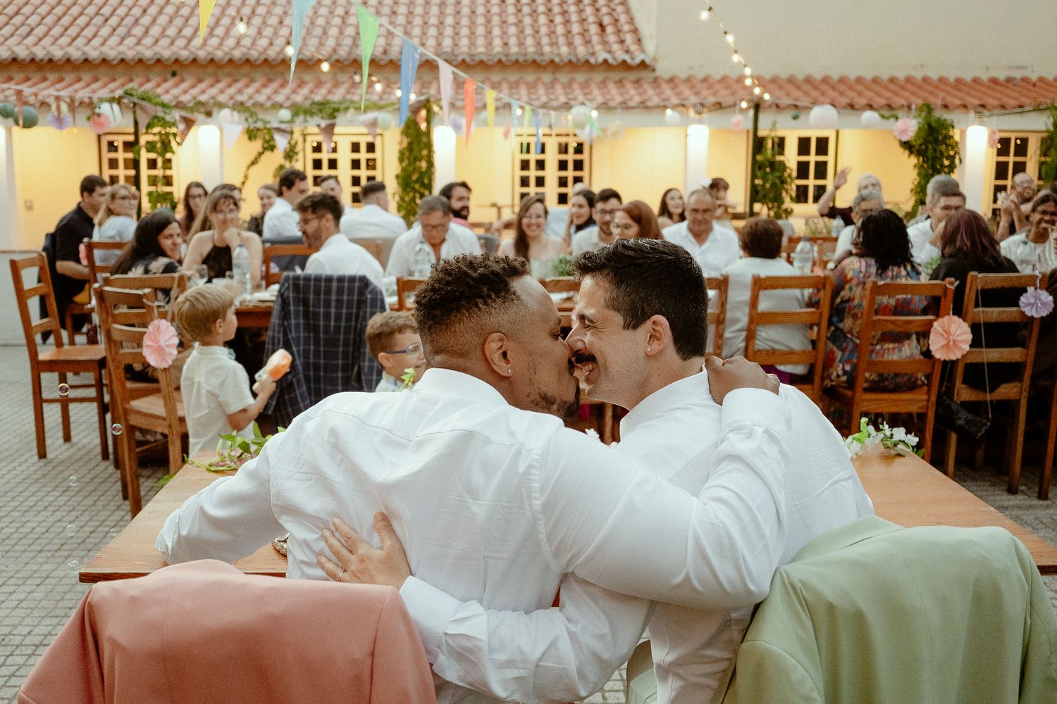 Two grooms sharing a kiss at their wedding reception with guests seated at decorated tables in background.