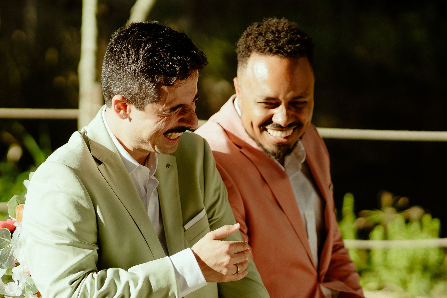 Two men in pastel suits laughing joyfully together outdoors at a wedding celebration.