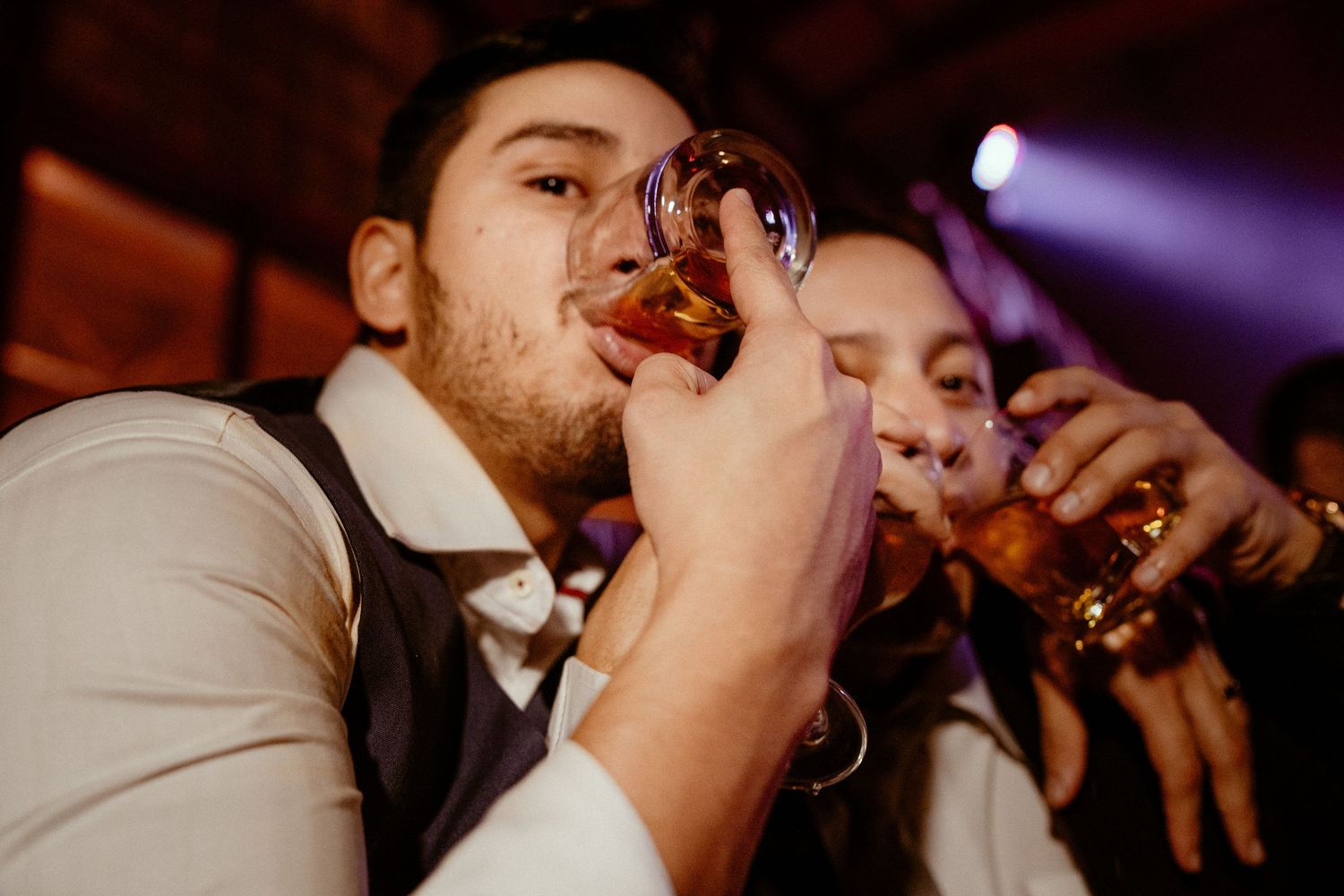 Shot glasses clink together in the air during a toast at a festive party celebration with purple lighting.