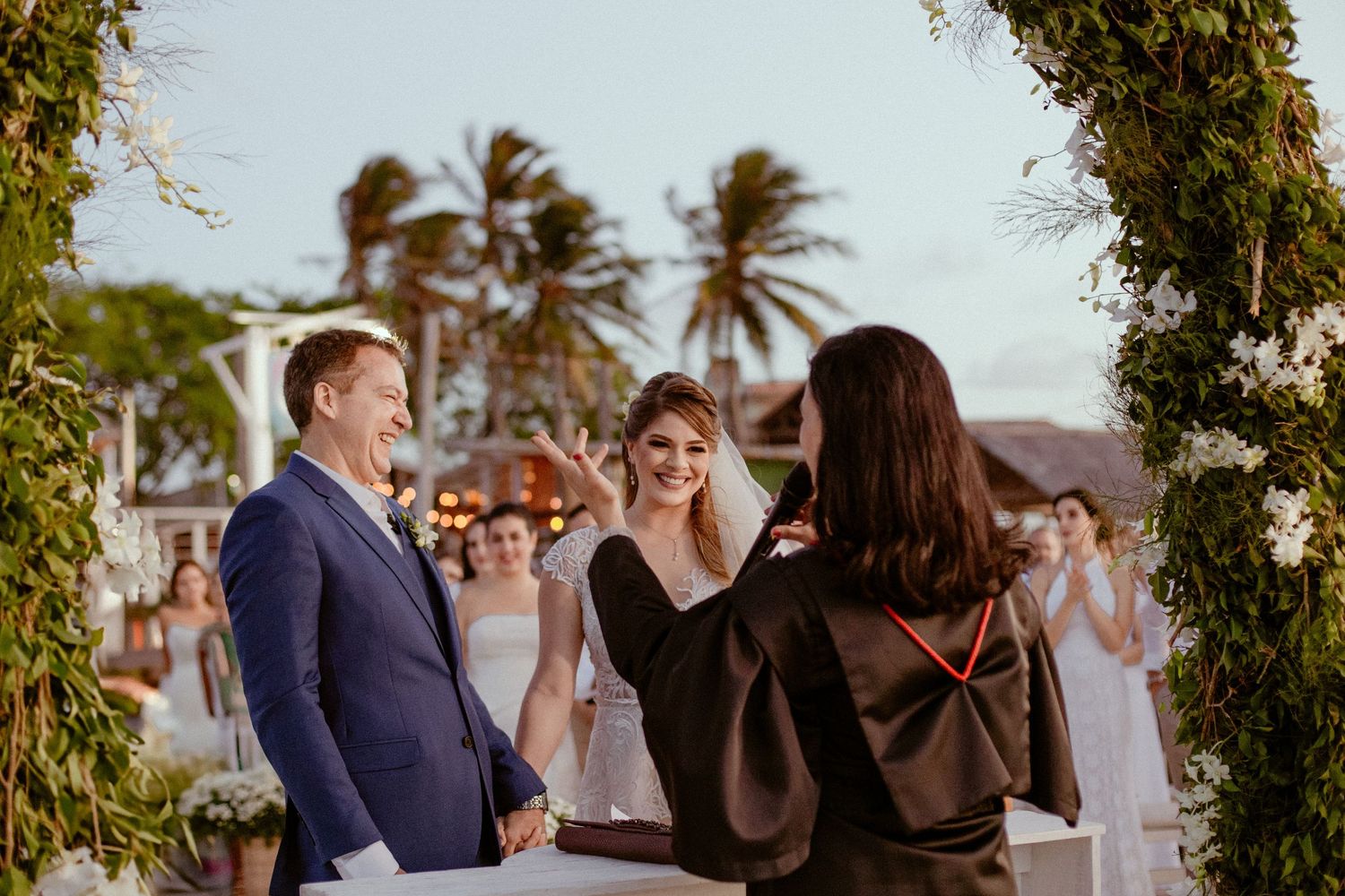 An outdoor wedding ceremony takes place under an arch with palm trees and tropical foliage in the background.