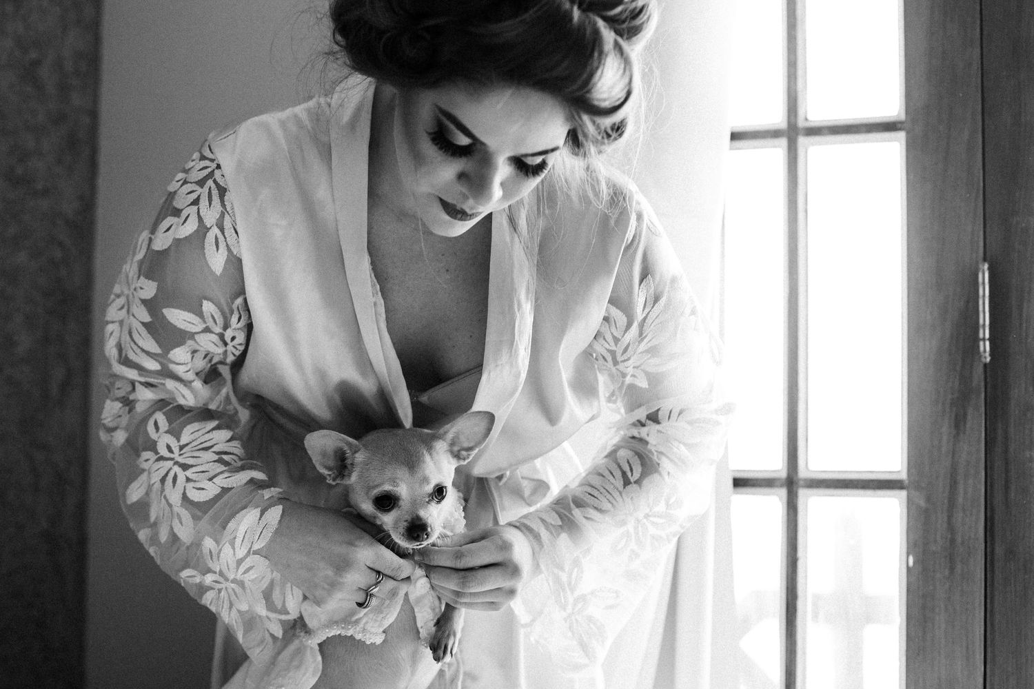 A romantic black and white wedding photograph with bride in ruffled dress leaning near window light.