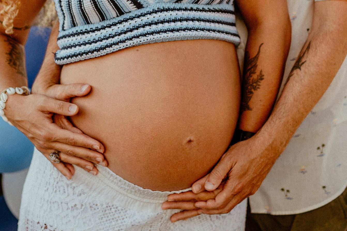 Close-up shot of hands gently cradling a pregnant belly, wearing a striped crop top and white lace shorts.