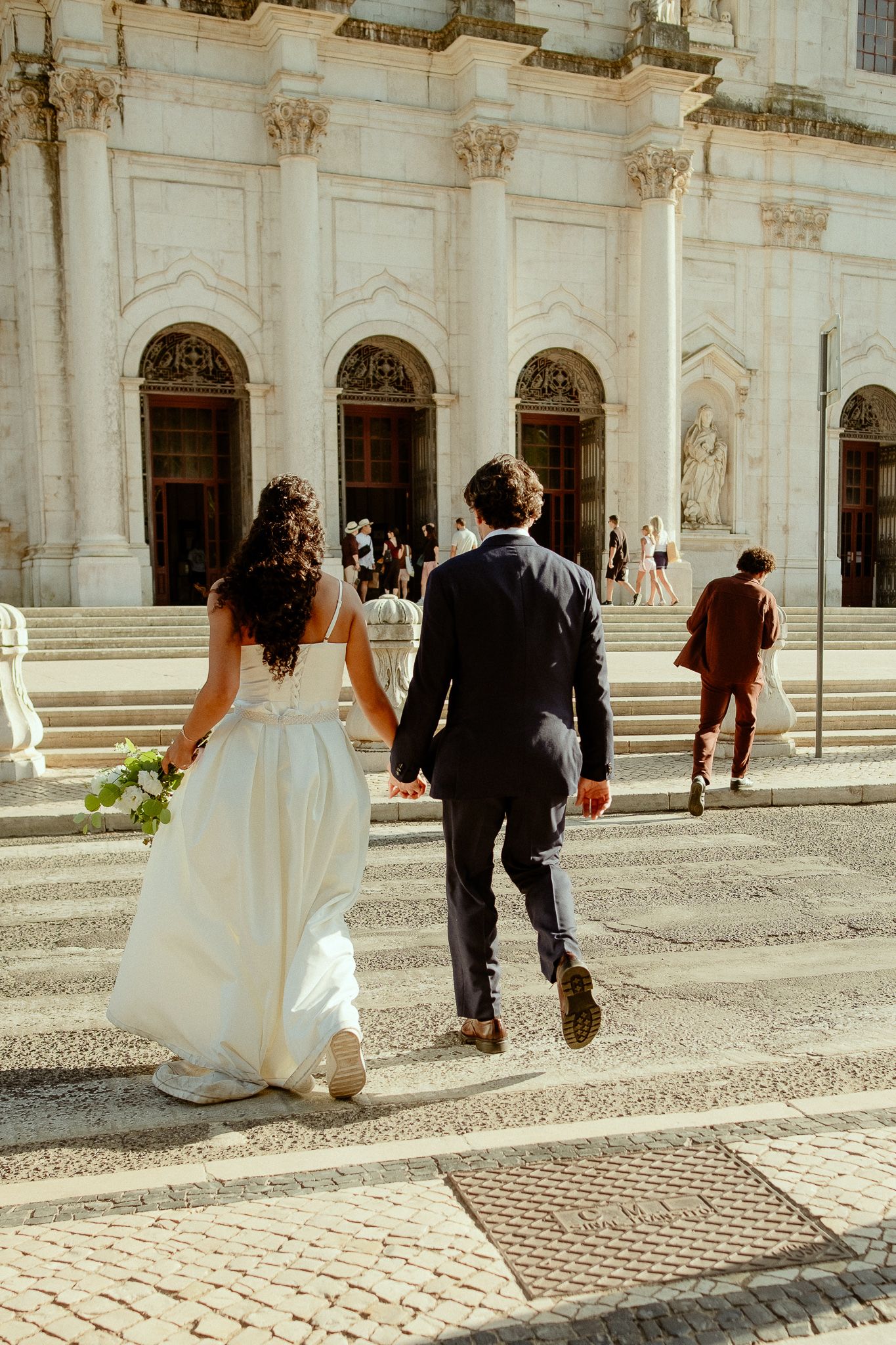 Bride and groom walking hand in hand toward a grand white classical building on their wedding day.