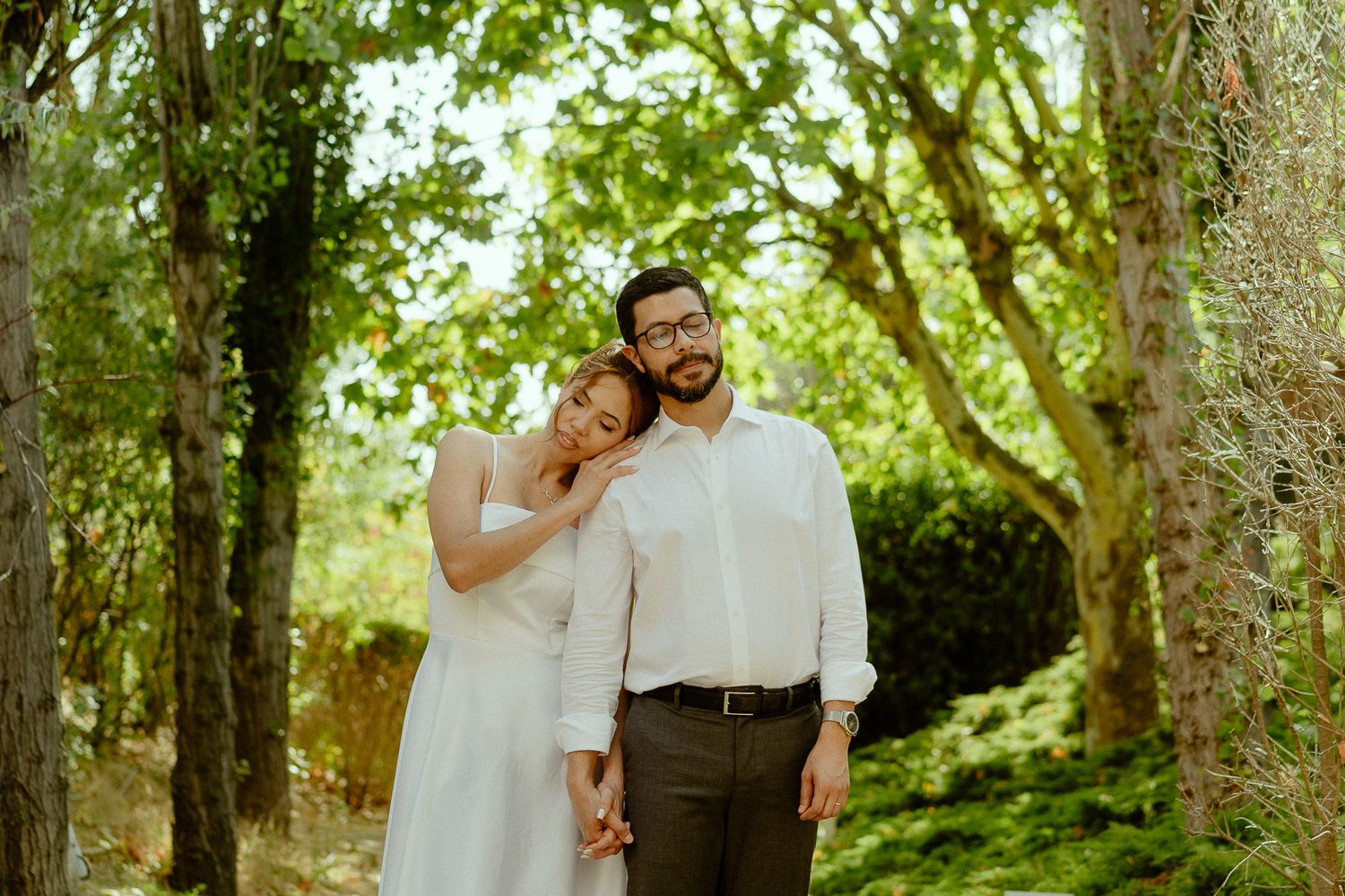 A couple poses in a lush green forest, woman in white dress leaning on mans shoulder, man wearing glasses and white shirt.
