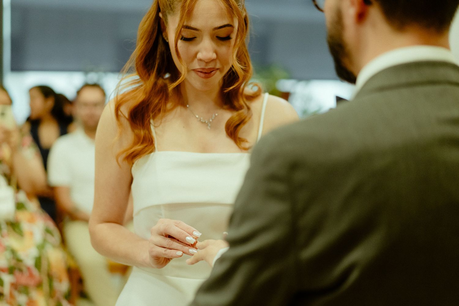Bride in white gown placing wedding ring on groom's finger during indoor ceremony.