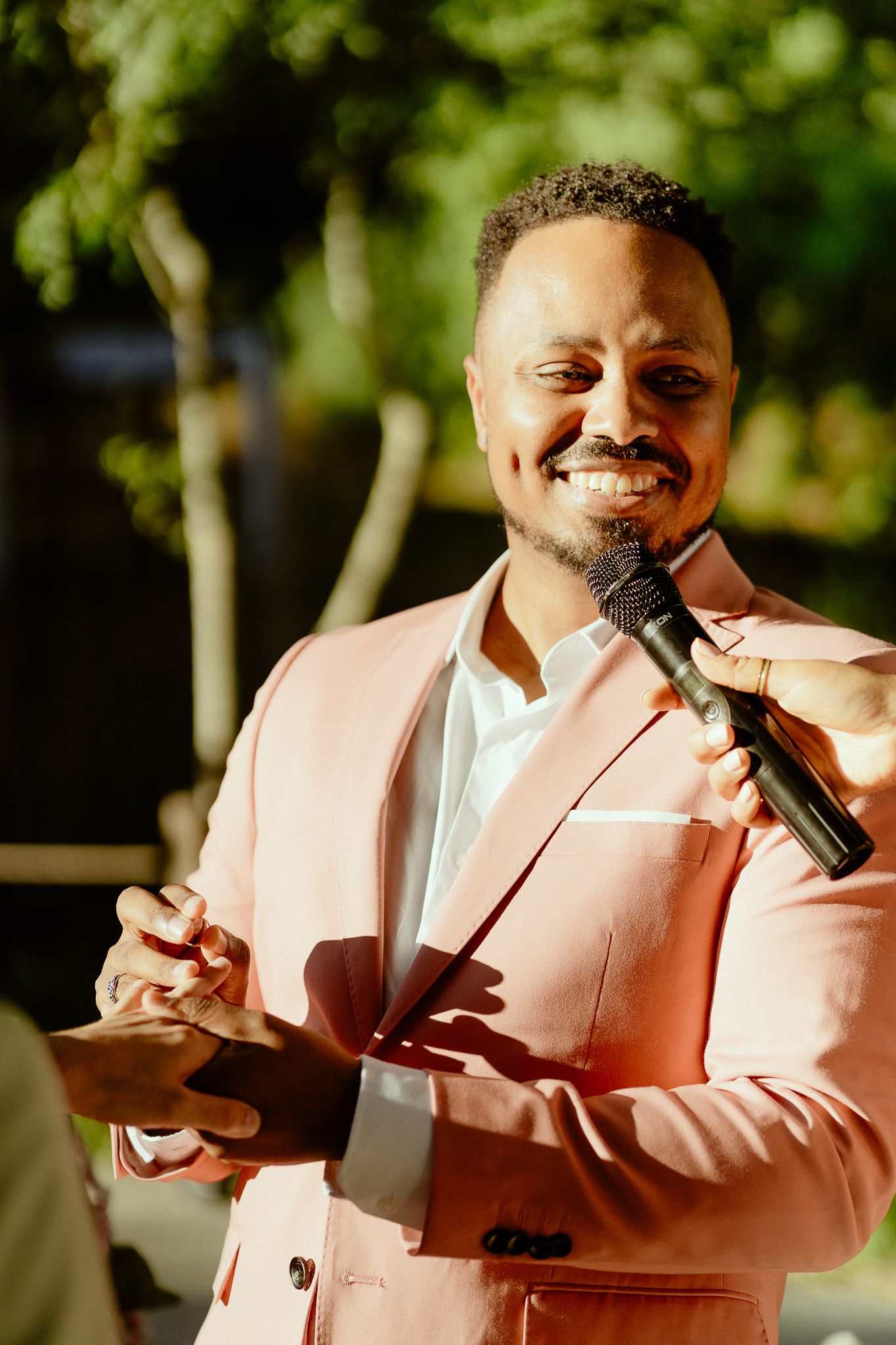 Smiling man in pink blazer holds microphone outdoors, speaking at an event with green trees in background.