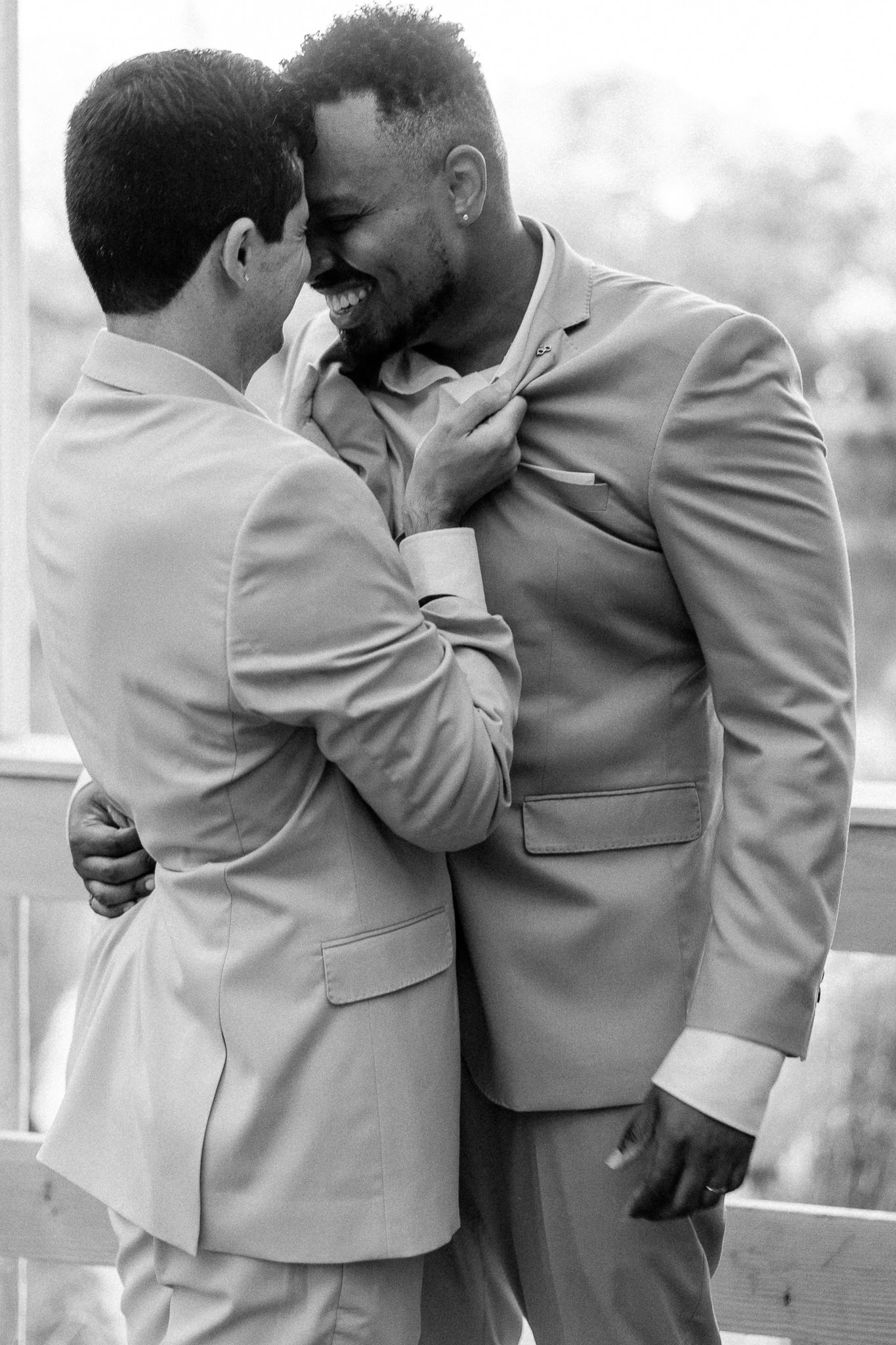Two grooms in matching suits share a joyful embrace, laughing together in a black and white wedding photo.