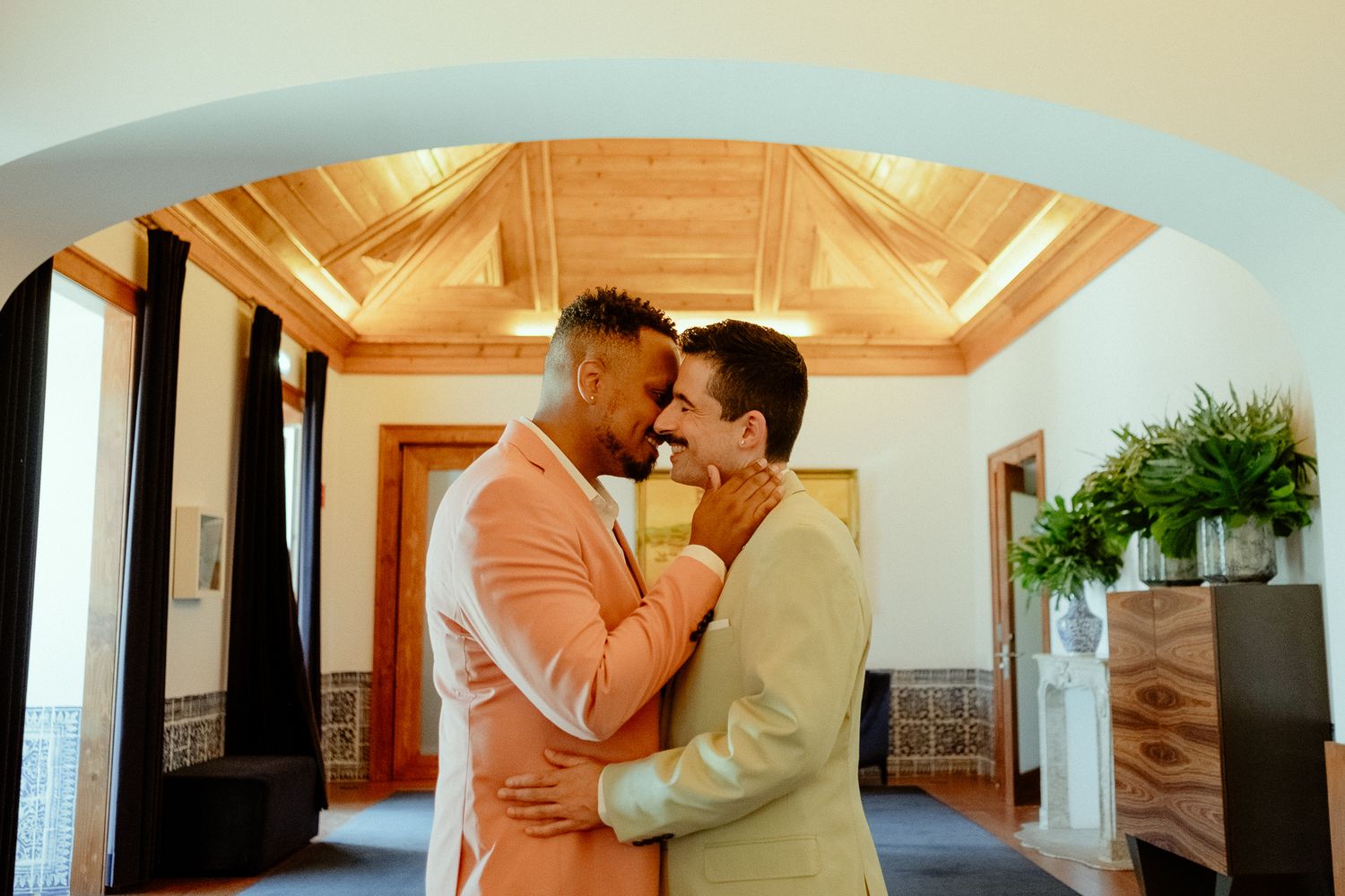 Two grooms in pastel suits share a romantic moment in an elegant arched venue on their wedding day.