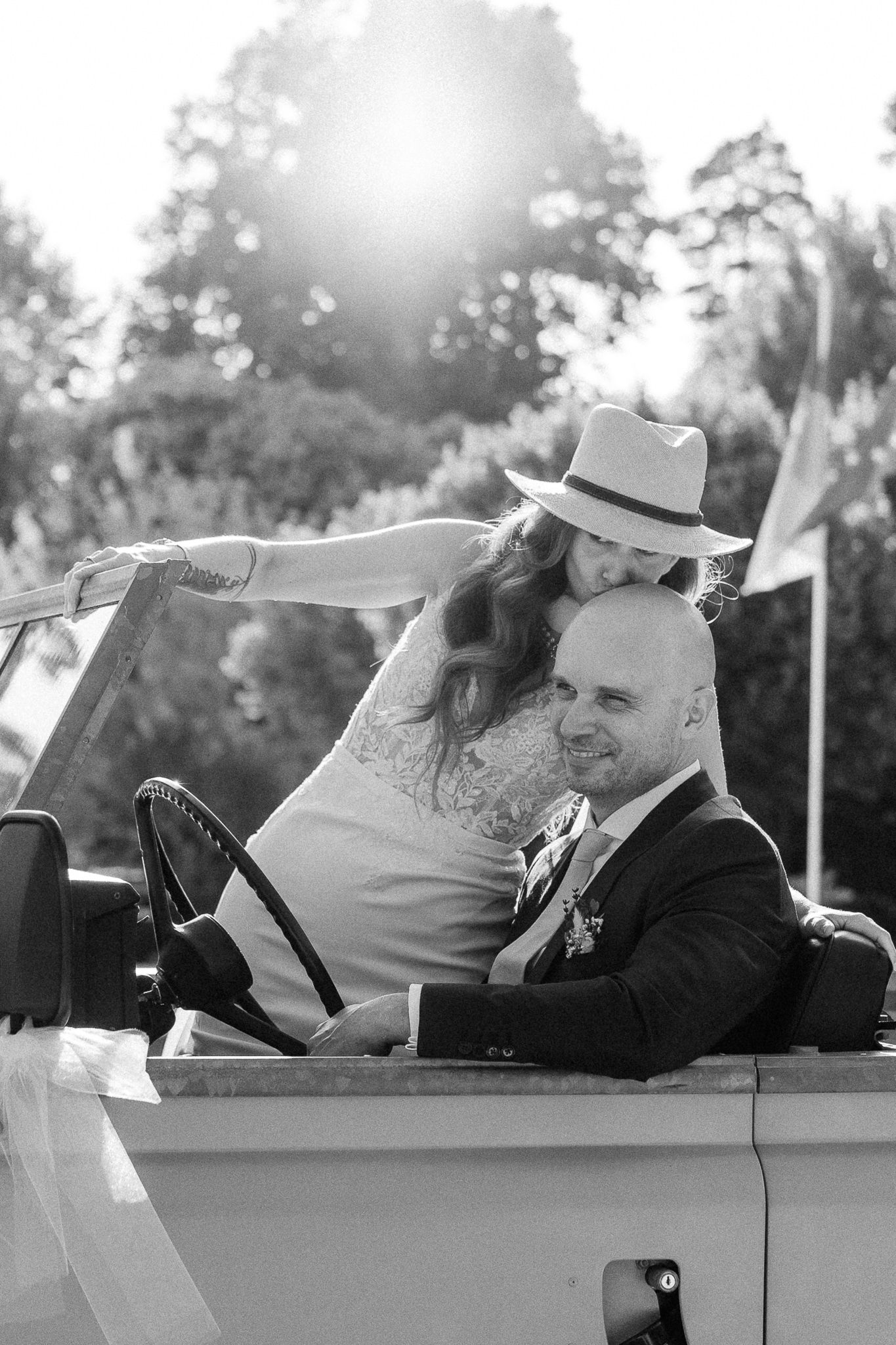 Black and white photo of a bride in a hat leaning over a groom sitting in a vintage convertible car.
