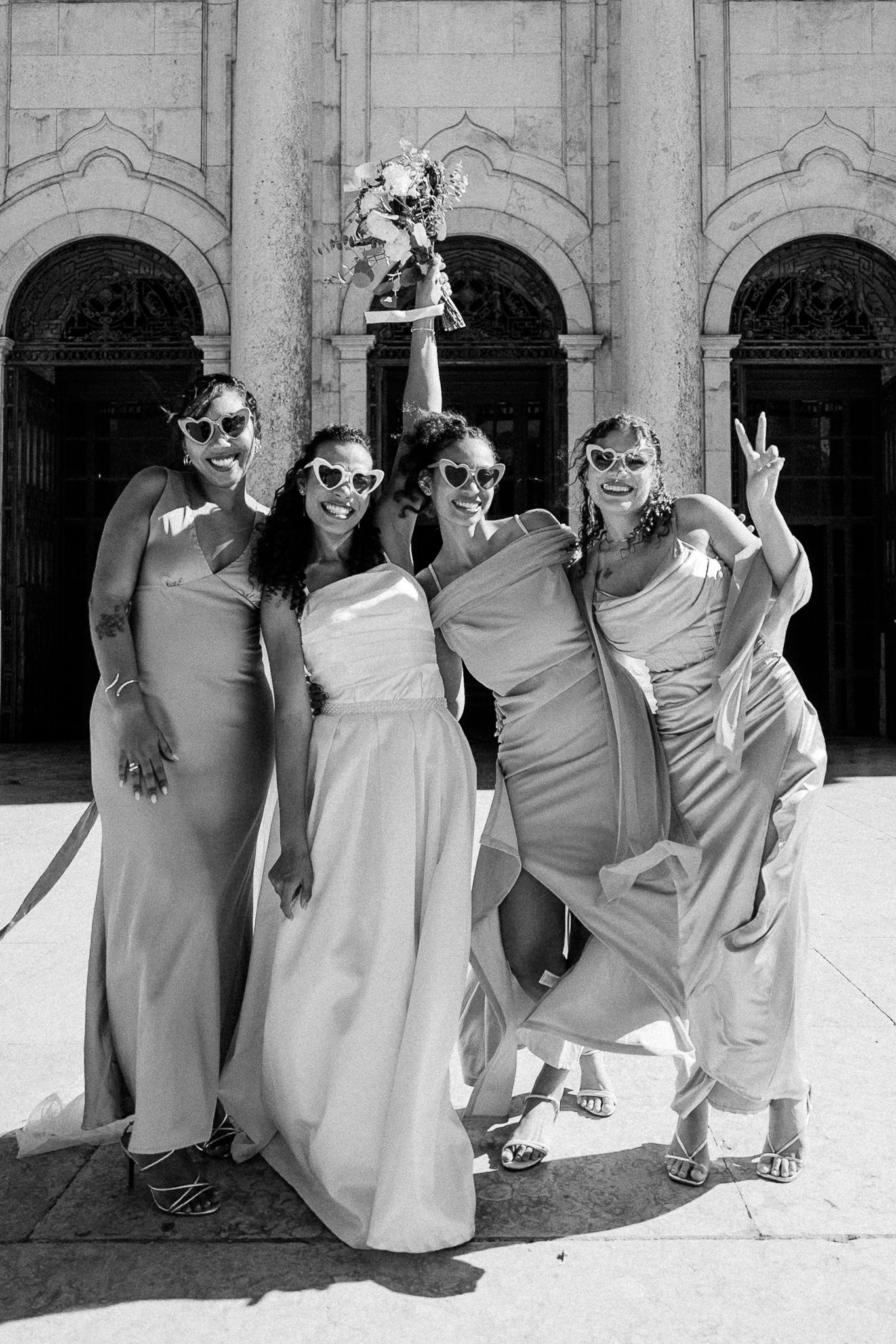 Black and white photo of a bride holding bouquet aloft with three bridesmaids in elegant gowns outside a grand building.