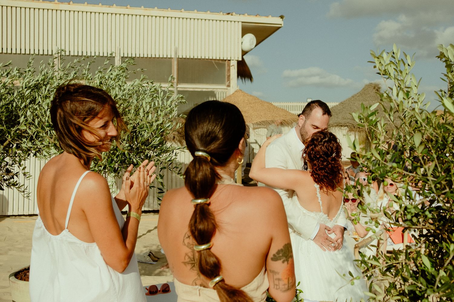 Bridesmaids in white dresses gather outdoors with bride and groom among olive trees at a sunny wedding celebration.