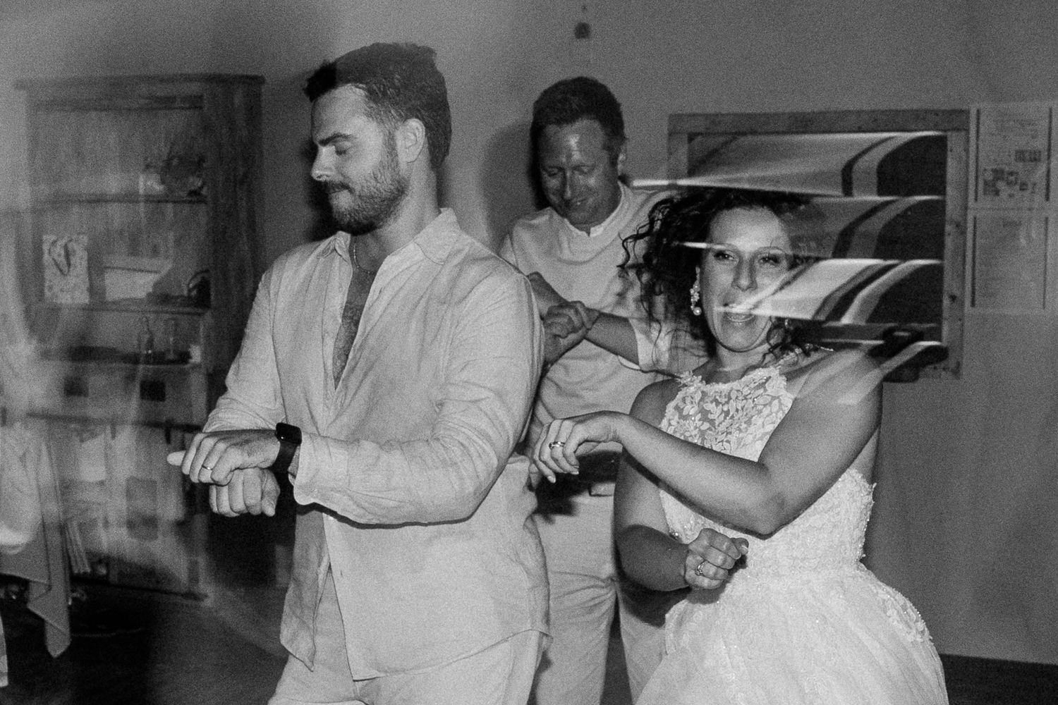 Black and white wedding photo of bride and groom dancing joyfully with guests at a vintage indoor reception.