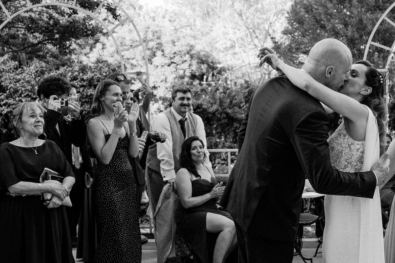 Black and white photo of a bride and groom sharing their first kiss at an outdoor wedding ceremony with guests applauding.