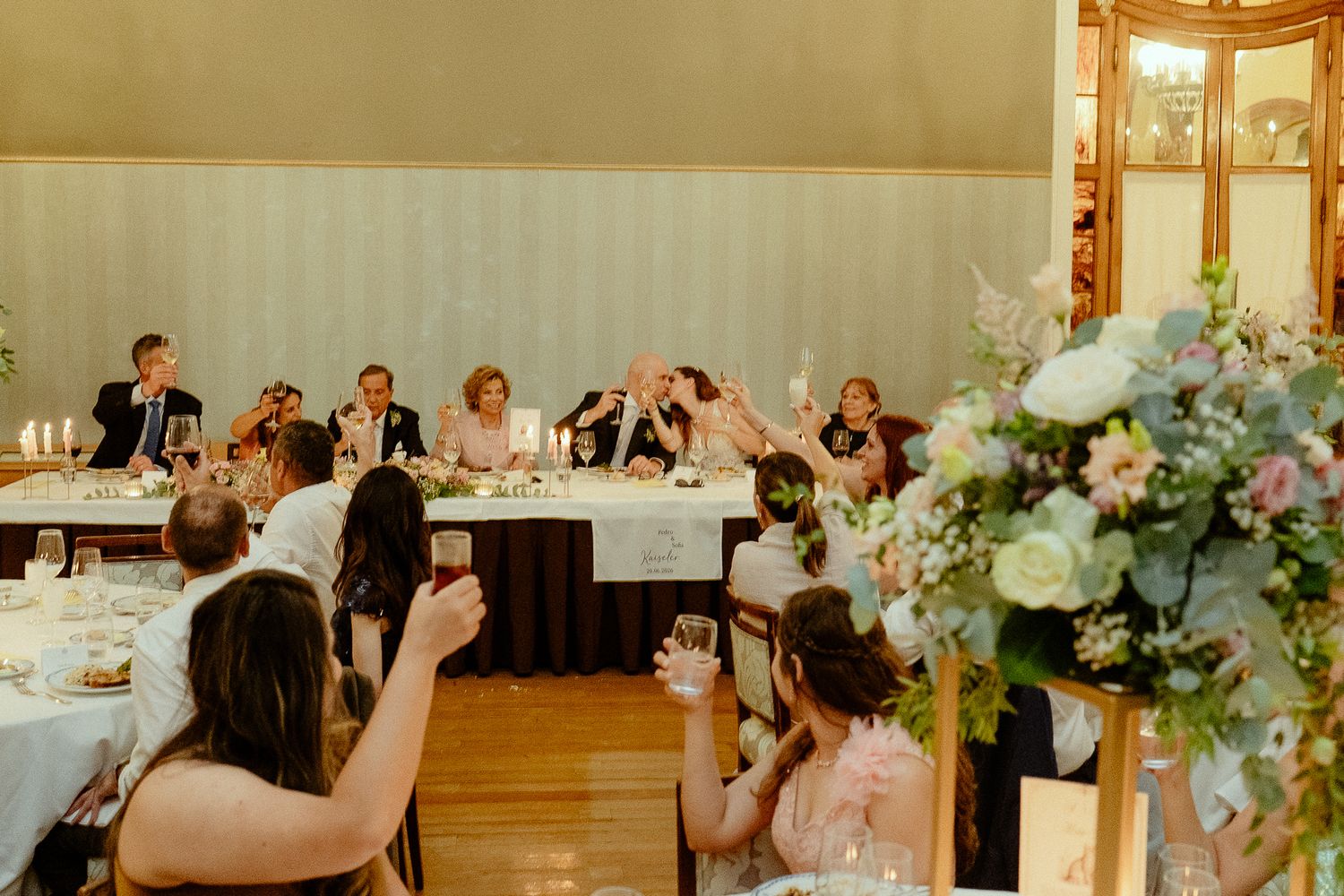 Guests raise glasses in a toast at an elegant wedding reception with floral centerpieces and a head table.