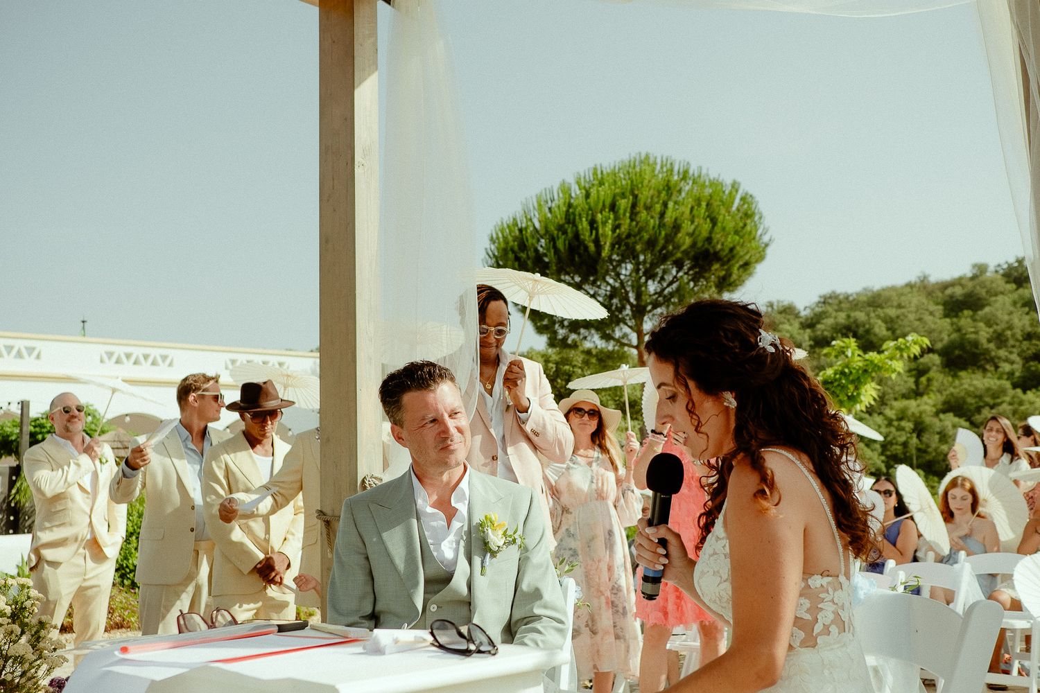 Groom seated at outdoor wedding ceremony table smiles as bride speaks into microphone under sunny pergola.