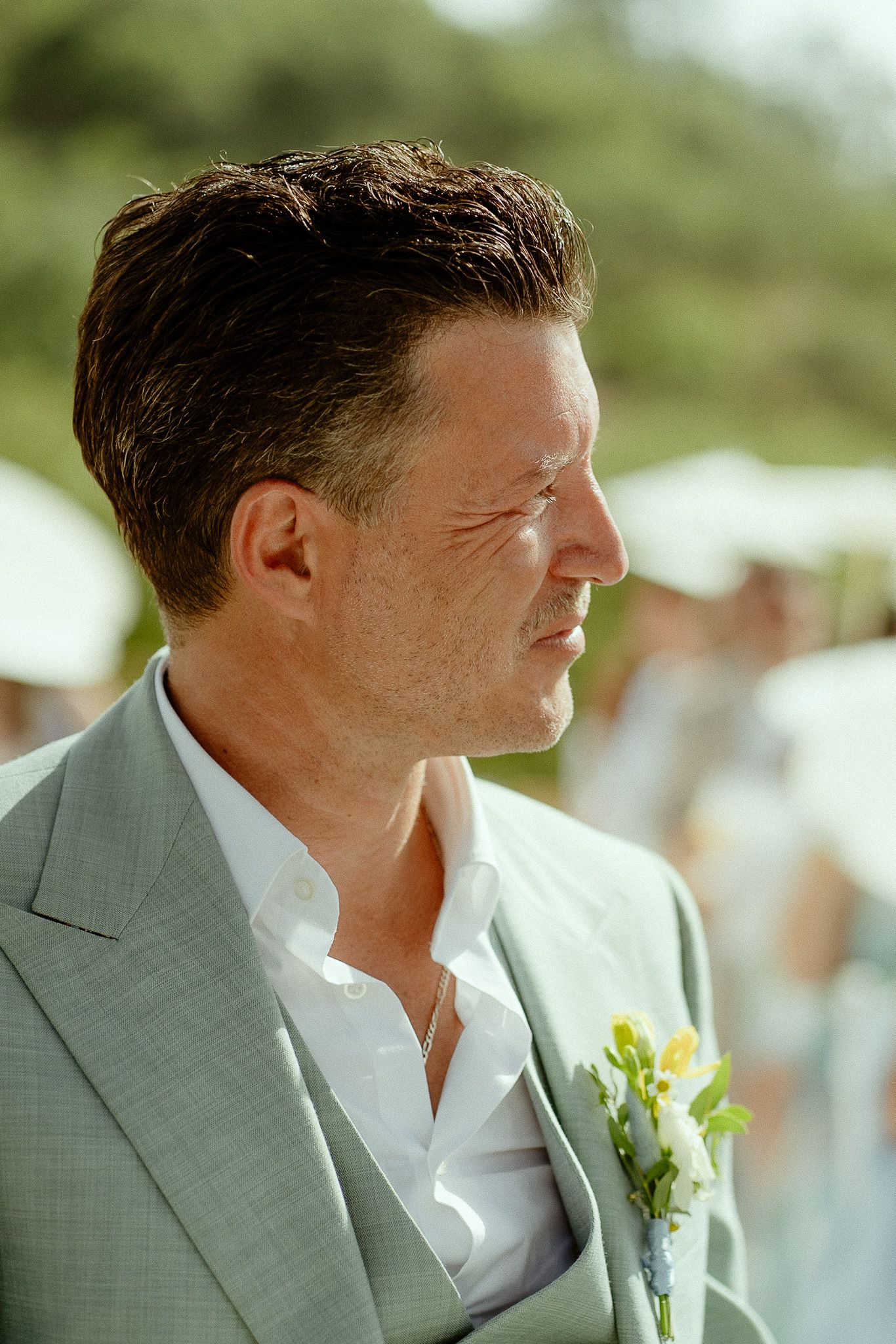 A groom in a sage green suit with a yellow floral boutonniere gazes thoughtfully at an outdoor wedding ceremony.