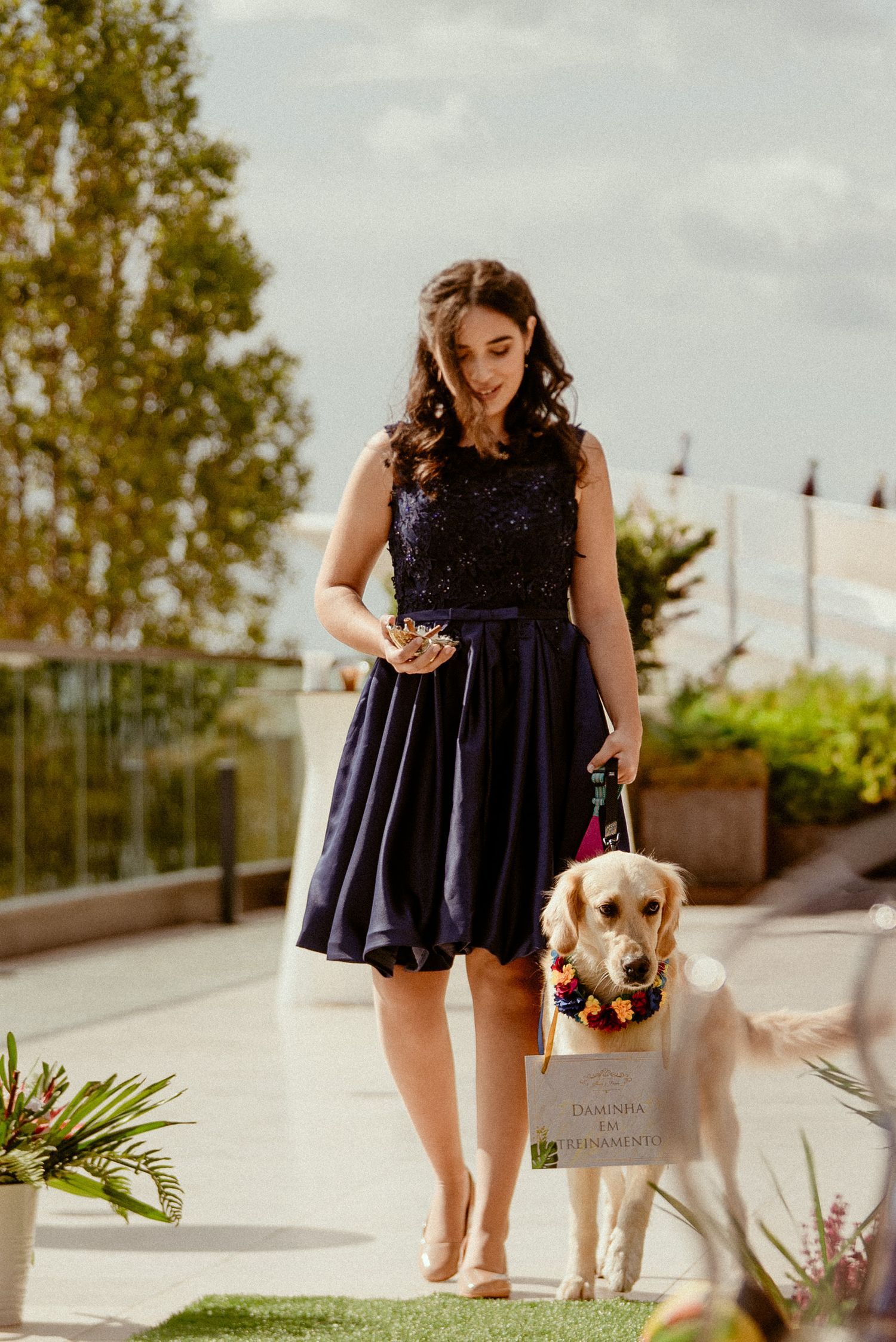 A person in a navy dress walks outdoors with a small dog on a sunny day in a landscaped setting.