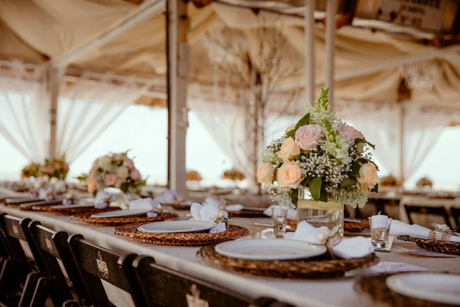 Elegant wedding reception tables set with rustic wooden chargers, white linens, and soft floral centerpieces in a bright venue.