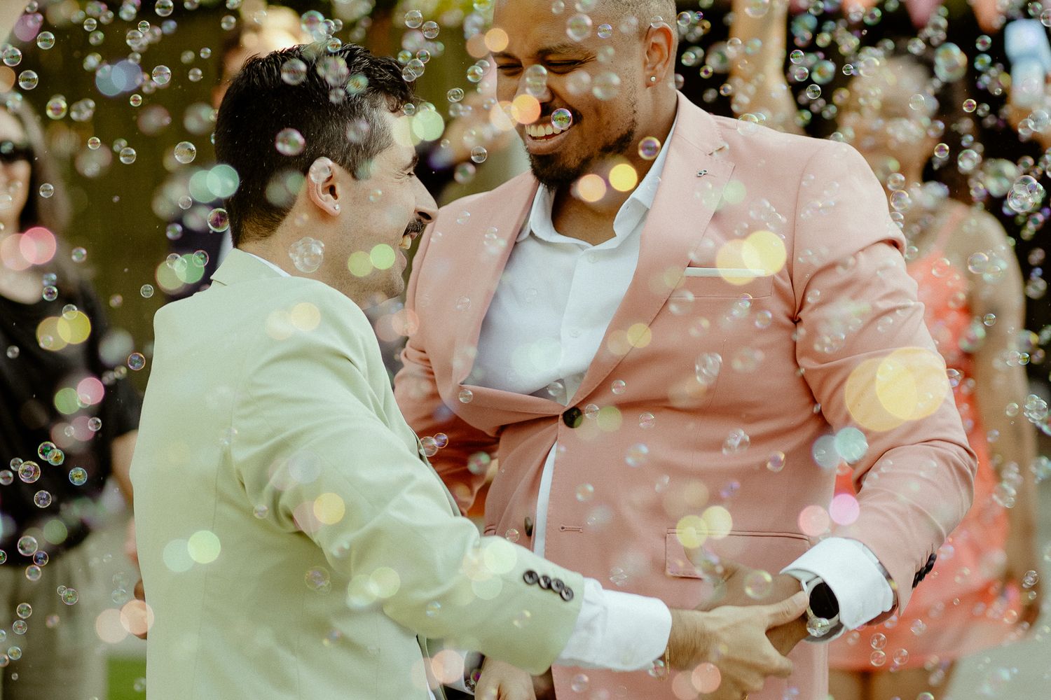 Two grooms in pink and white suits smiling and holding hands surrounded by bubbles at their wedding celebration.