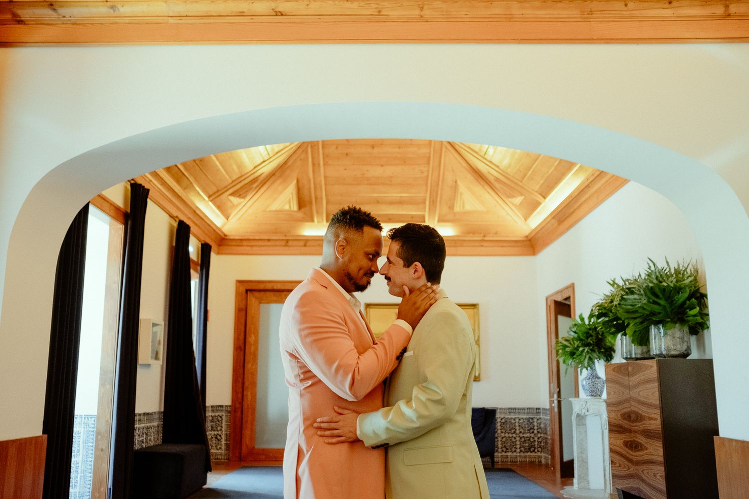 Two grooms share a tender embrace under an arched doorway in an elegantly decorated wedding venue.