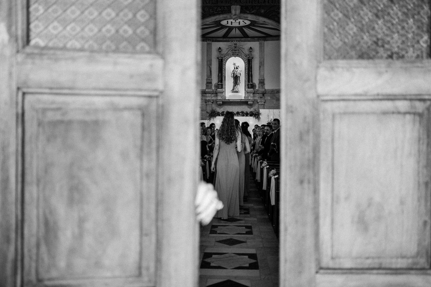 Black and white photo of a woman walking down a church aisle viewed through ornate wooden doors.