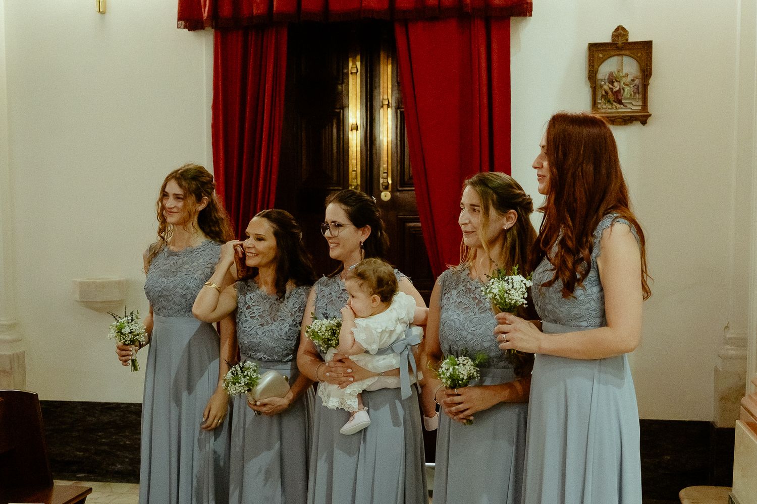 Five bridesmaids in matching gray dresses holding white bouquets stand together in a church, one holding a baby.