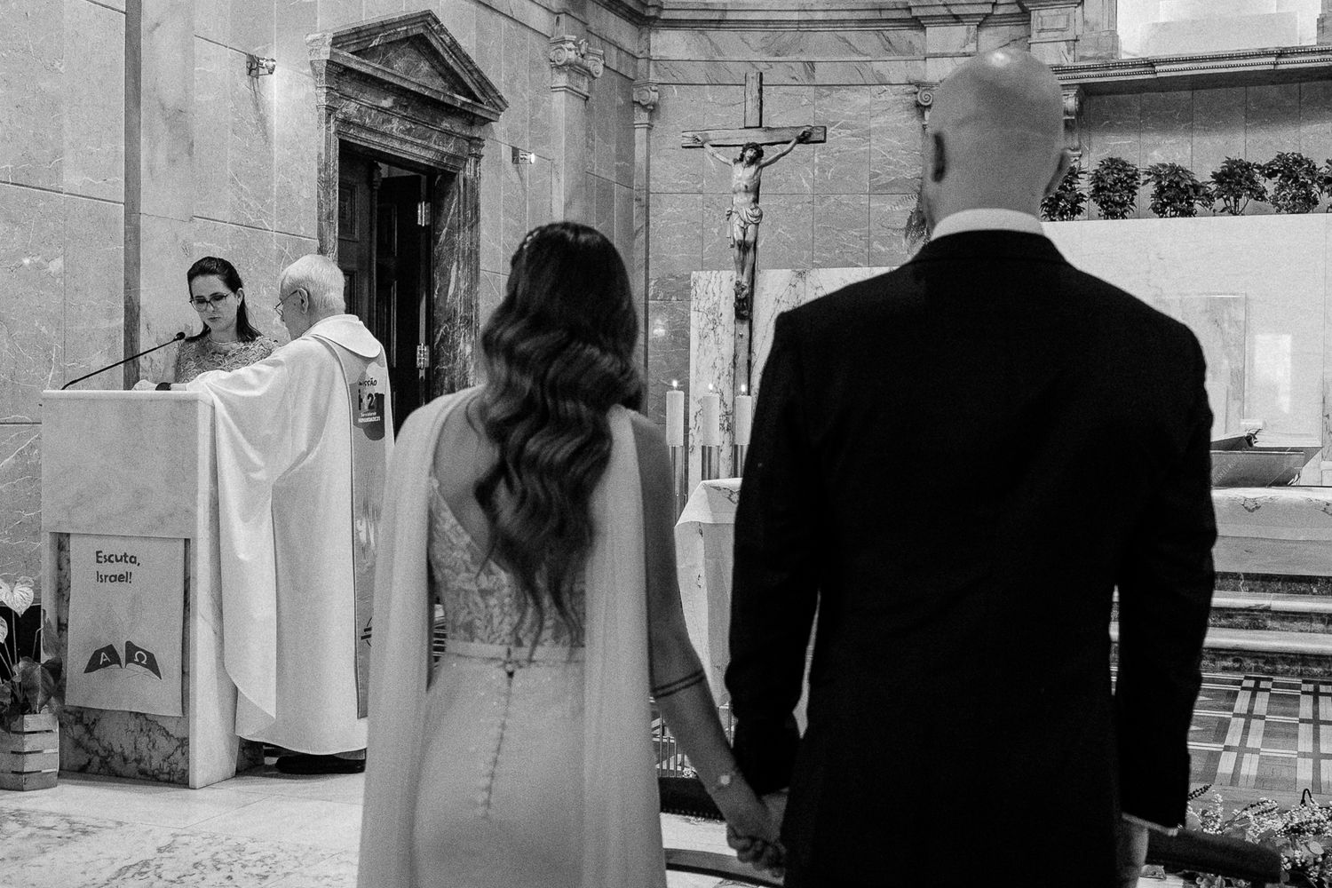 Black and white wedding ceremony photo of couple holding hands before priest at church altar with crucifix.