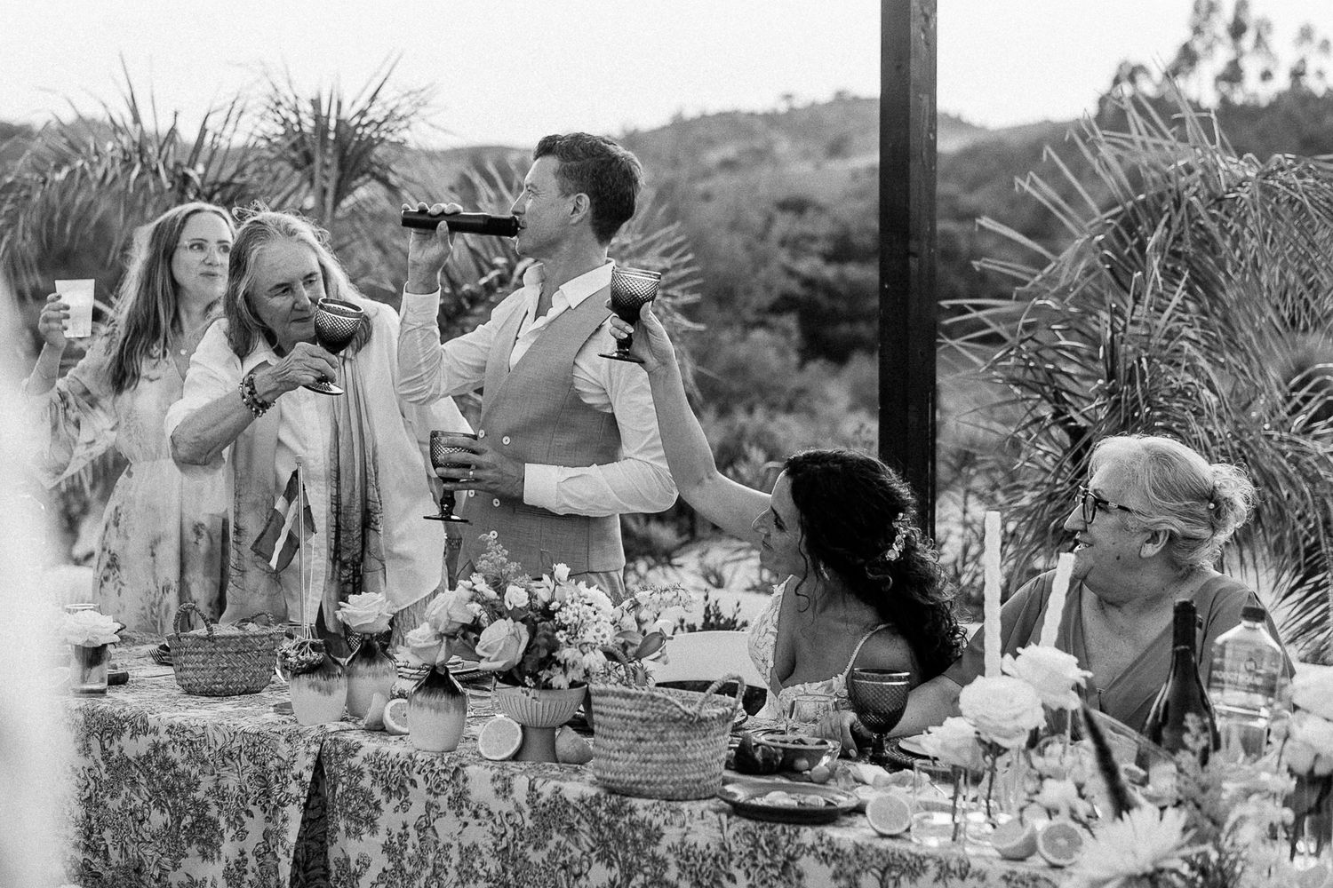 Black and white wedding reception photo showing guests toasting at an outdoor table with floral arrangements and scenic hills.
