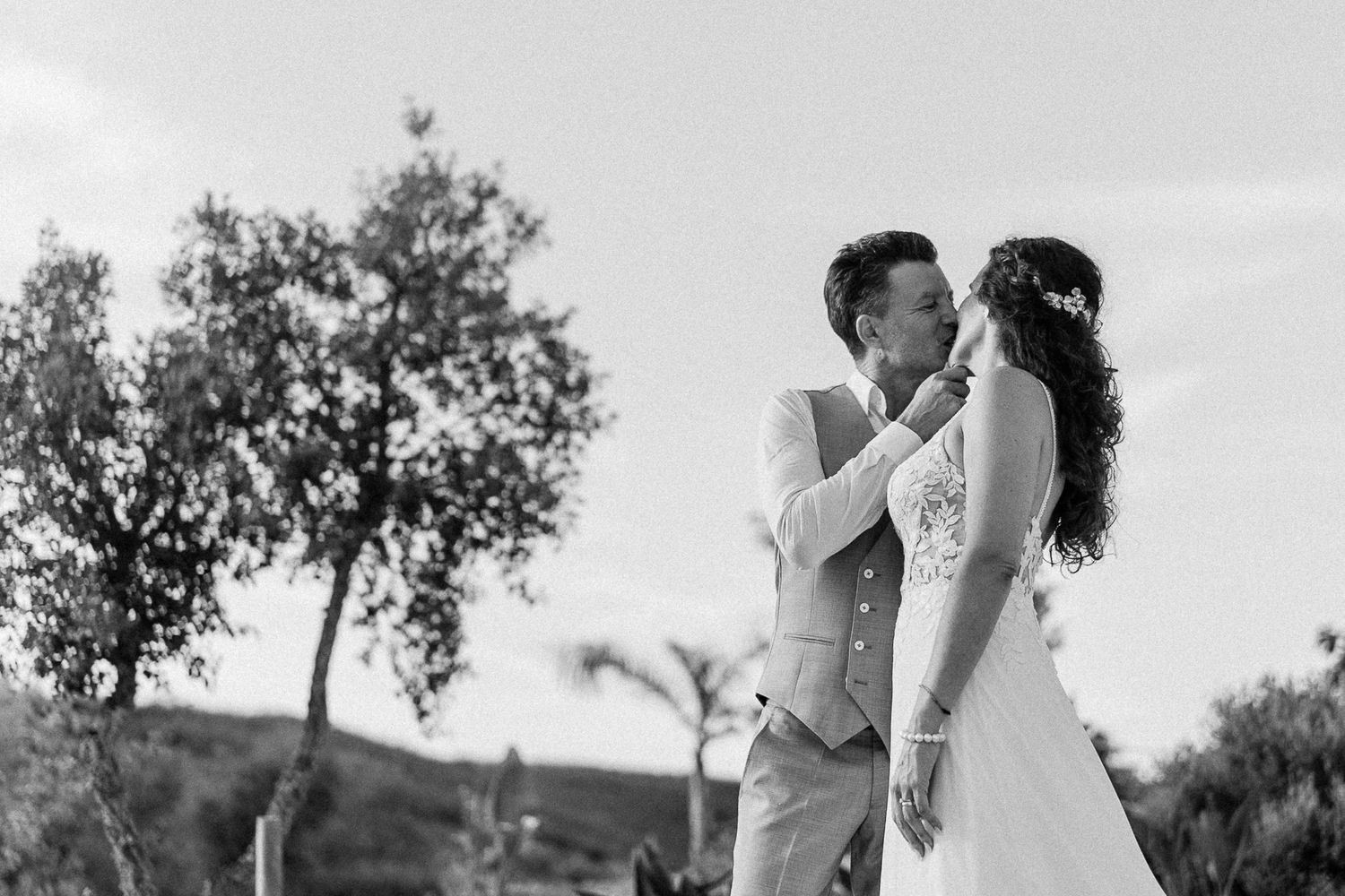Black and white wedding photo of a couple sharing a romantic moment outdoors with trees and hills in the background.