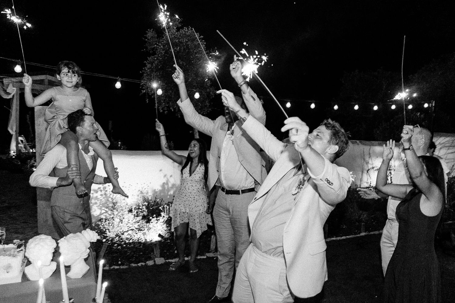 Black and white photo of wedding guests celebrating outdoors at night with sparklers under string lights.
